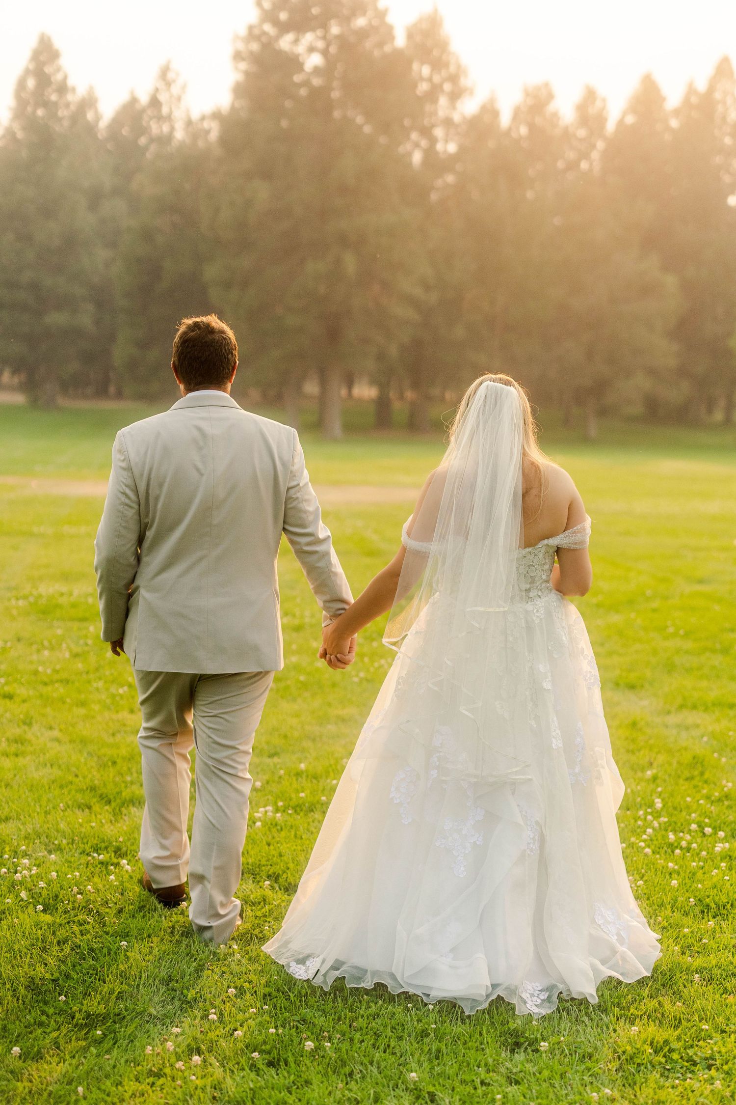 Bride and groom holding hands walking across lush green grass at sunset during wedding portraits.