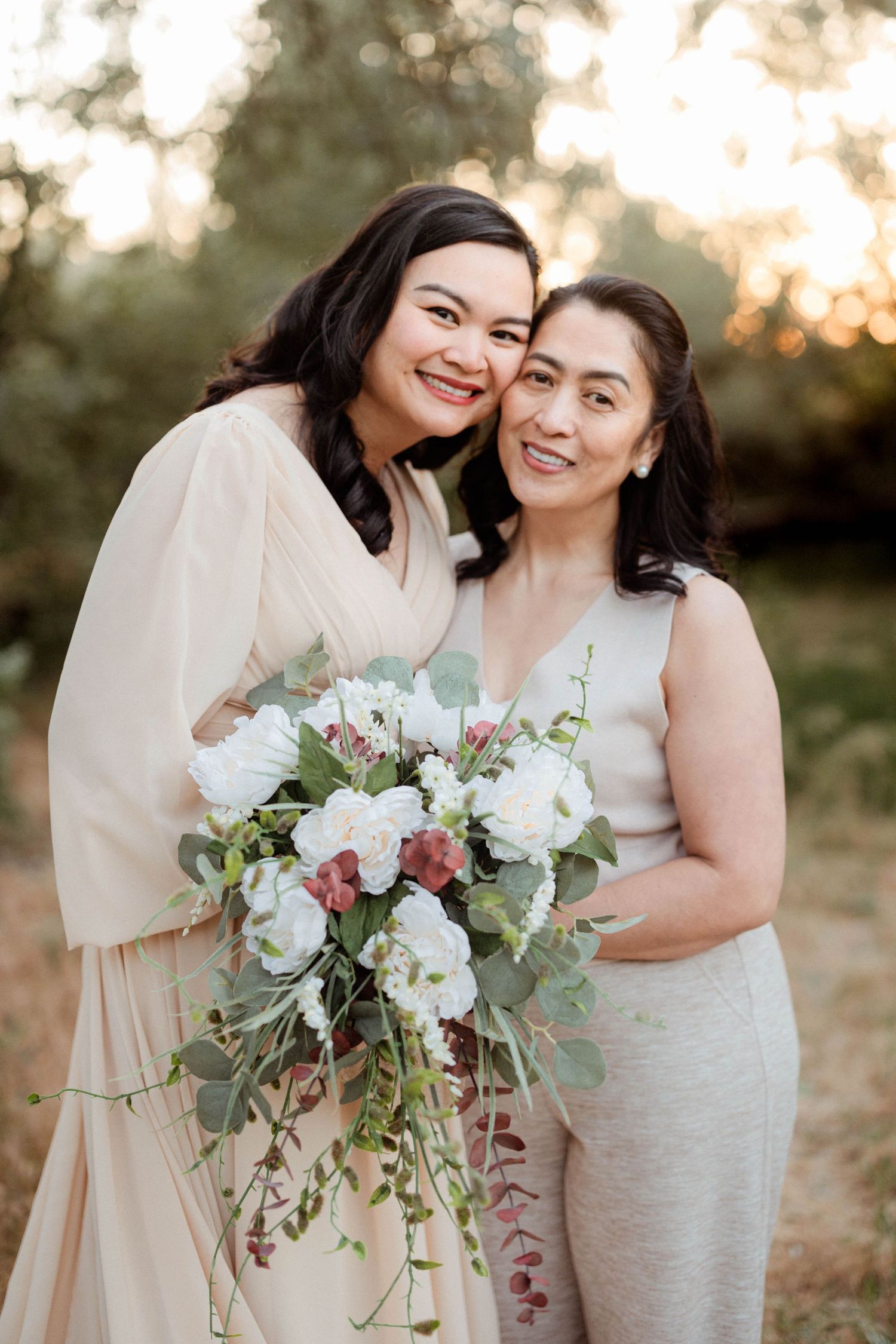 Two smiling women pose together outdoors, one holding a bouquet of white roses and greenery.