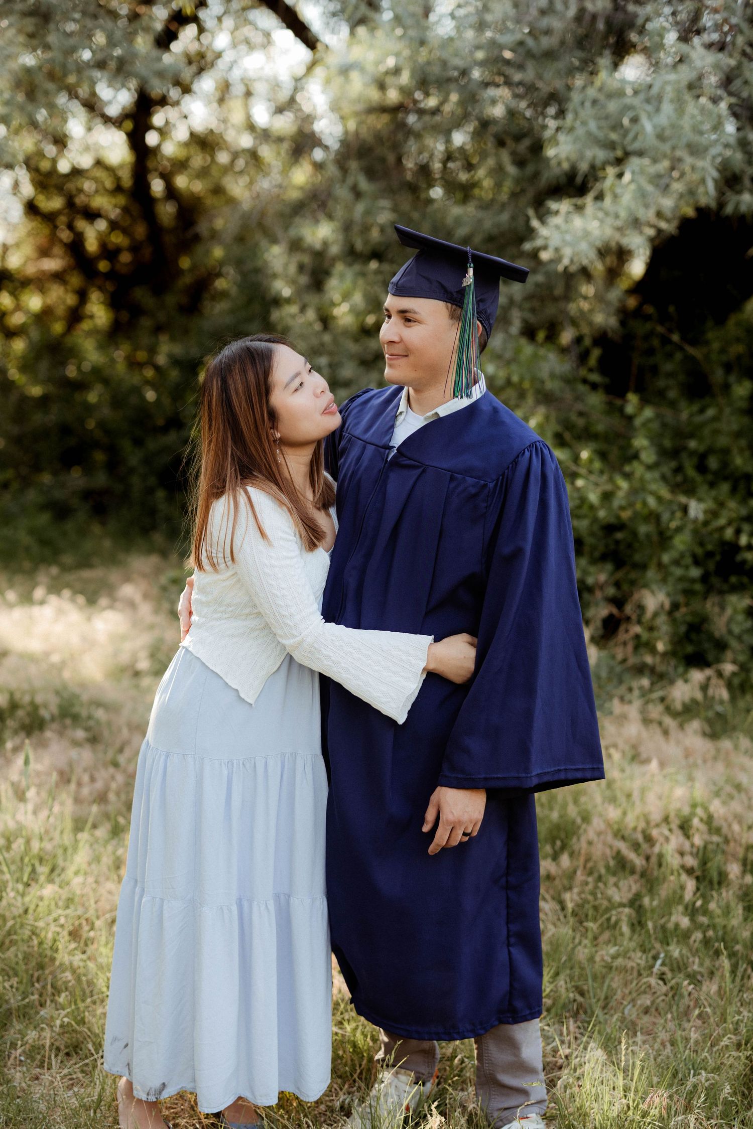 A young woman embraces a graduate in cap and gown outdoors among green trees.