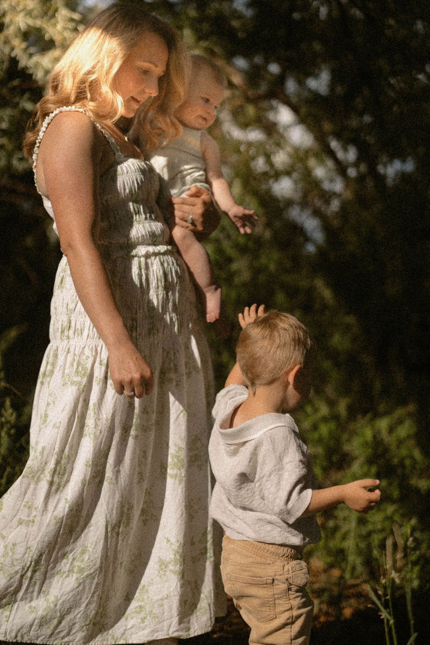 A mother in a white floral dress holds her baby while her young son stands beside her in dappled sunlight.