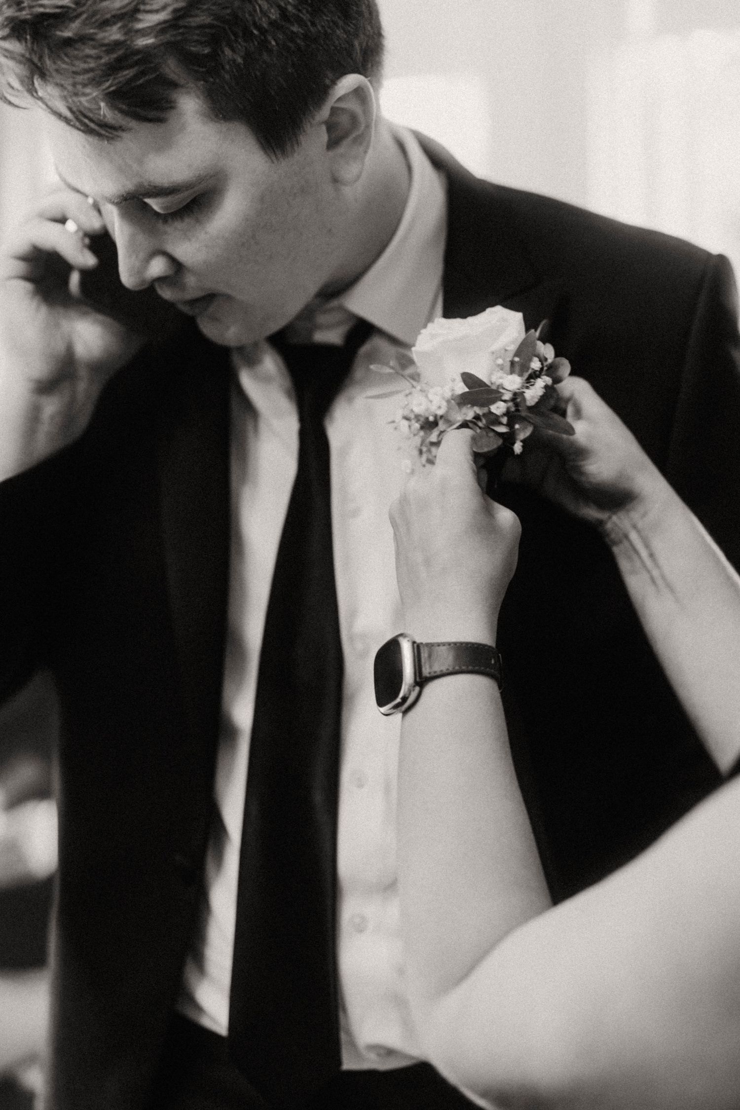 Black and white photo of a boutonniere being pinned onto a groom's lapel before the wedding.