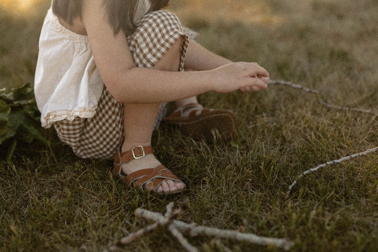 A child in a gingham outfit and brown sandals crouches on grass, playing with small sticks.