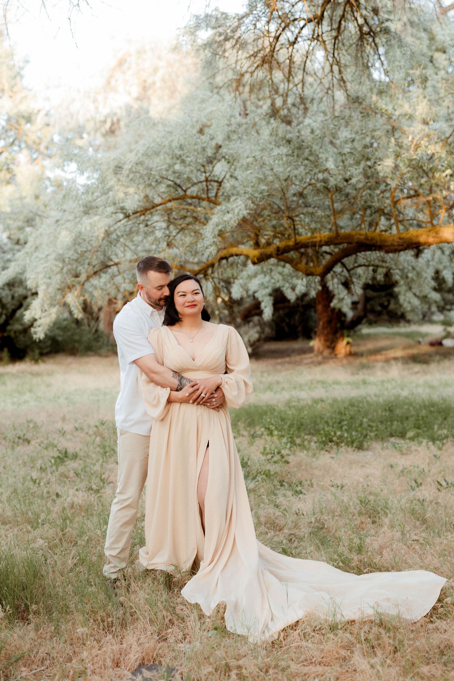 Man embraces woman from behind in flowing champagne dress during golden hour outdoor photo session.
