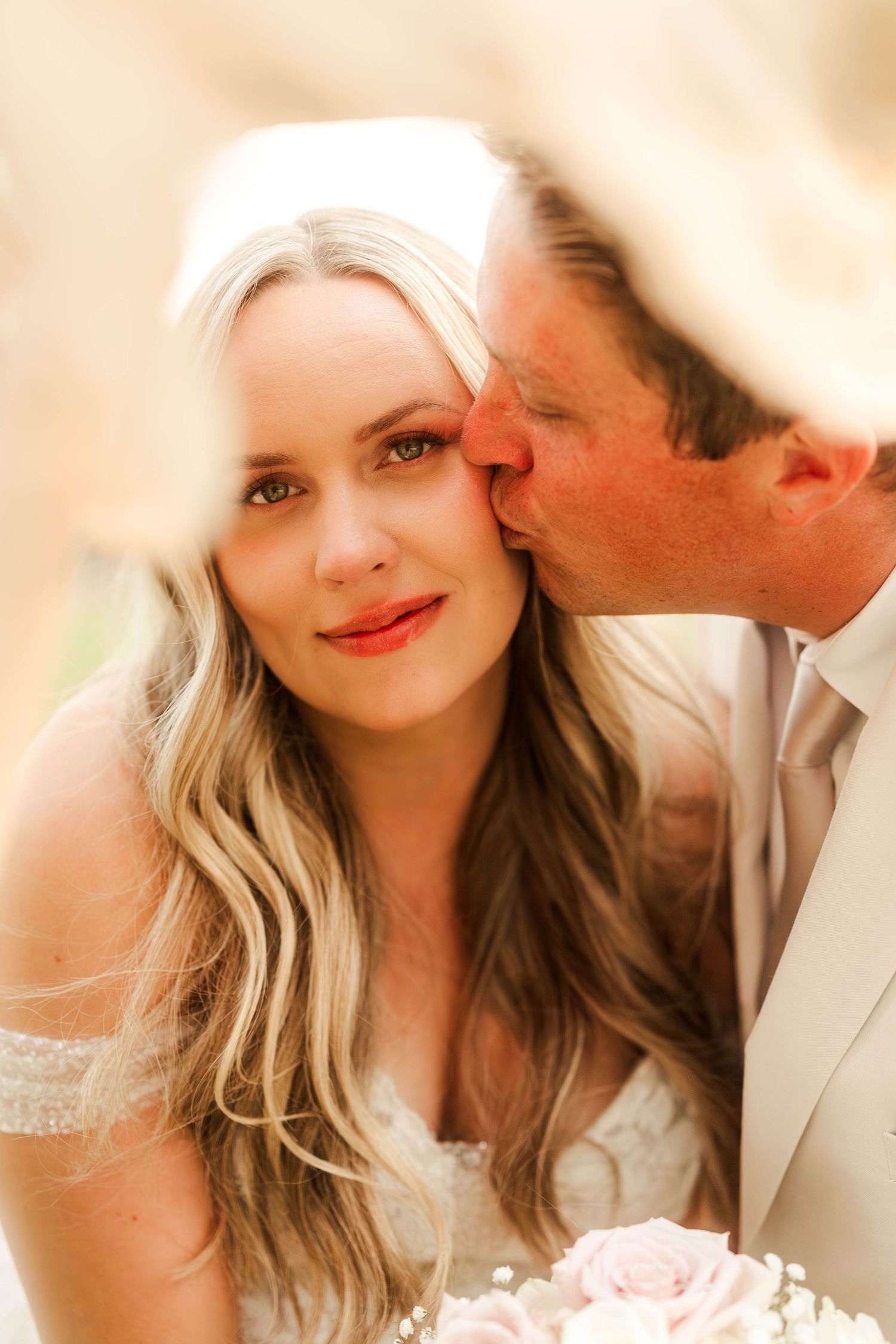 Groom tenderly kissing bride on the cheek as she smiles holding a bouquet at outdoor wedding.