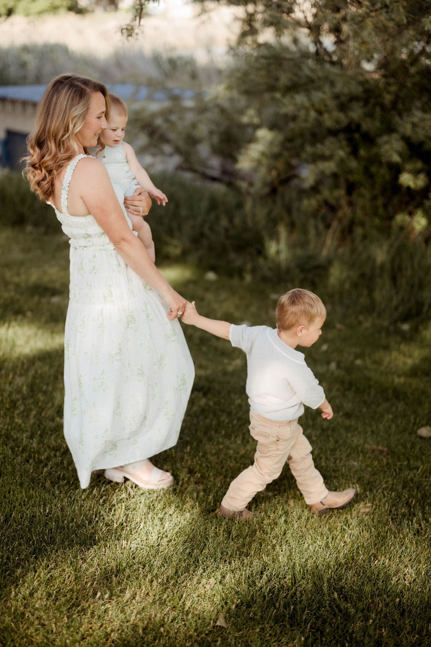 A mother in a white floral dress holds her baby while walking hand-in-hand with her toddler son outdoors.
