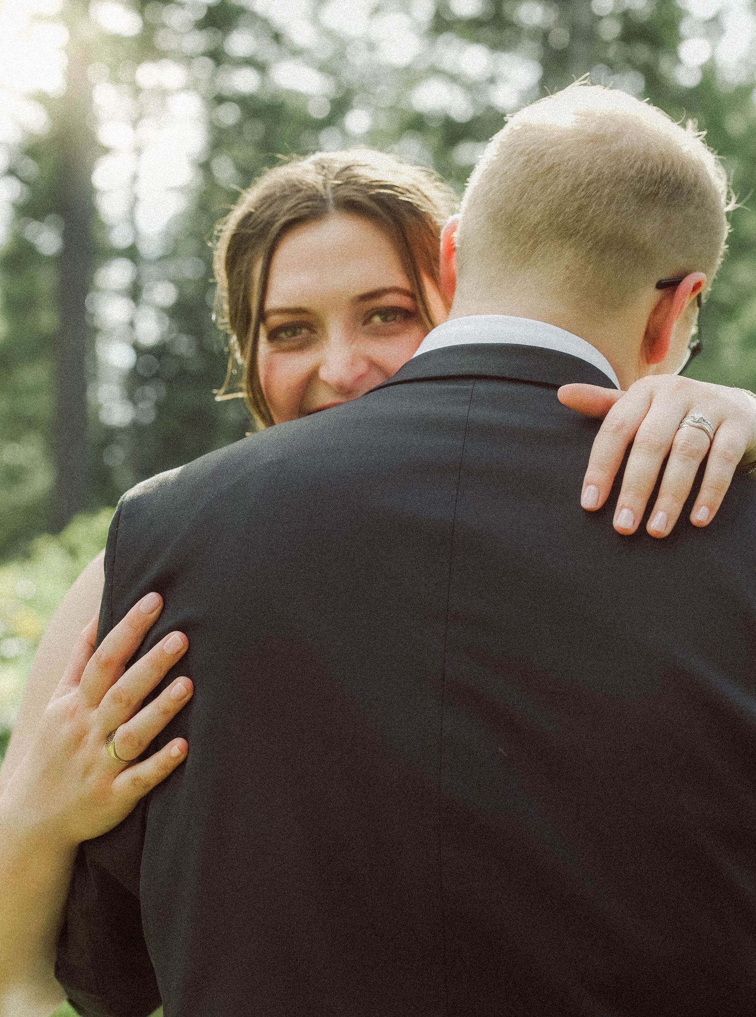 Smiling bride peeks over groom's shoulder while embracing him, showing her wedding ring, in a forest setting.
