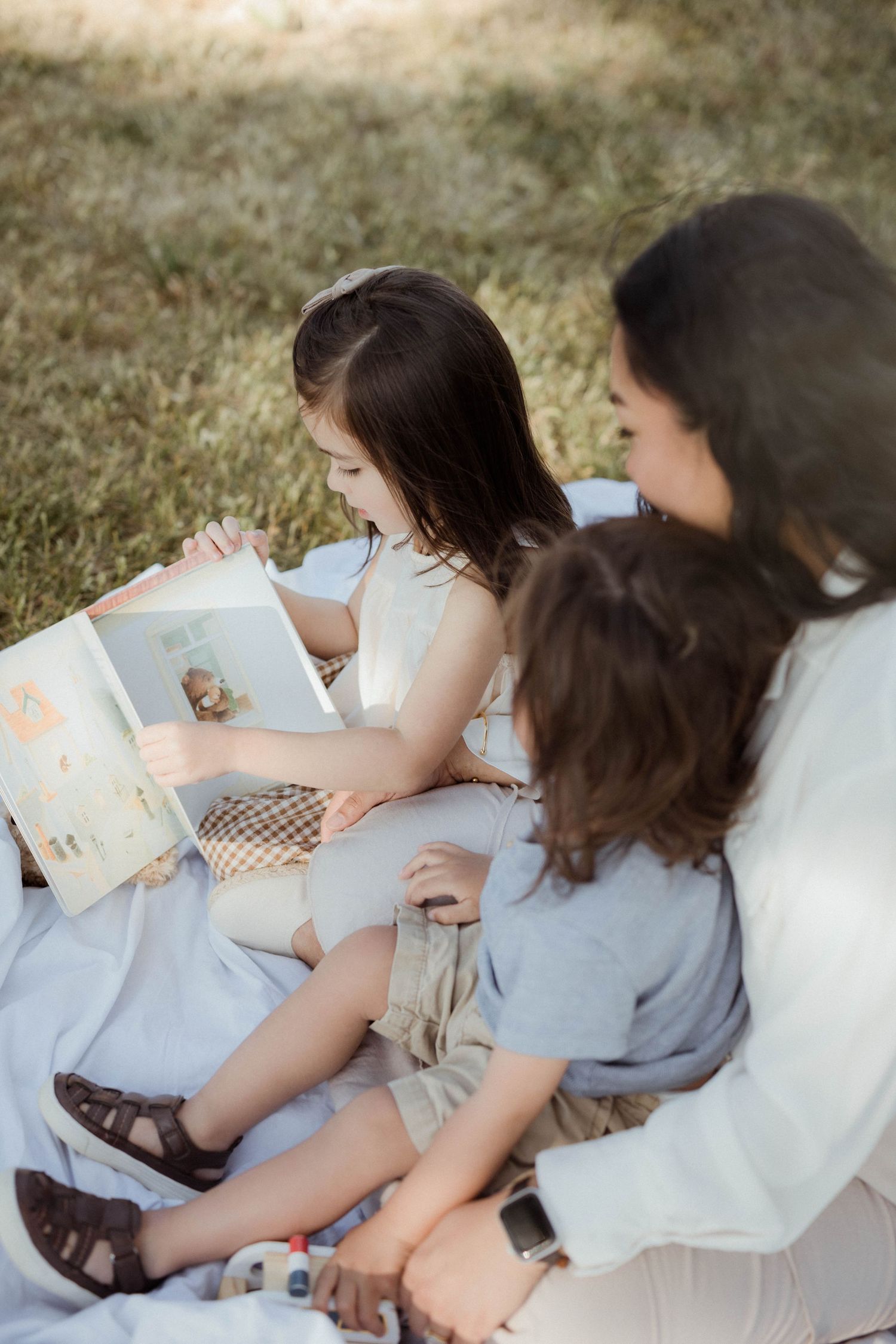 A mother and two young children sit on a blanket outdoors, reading a colorful picture book together.