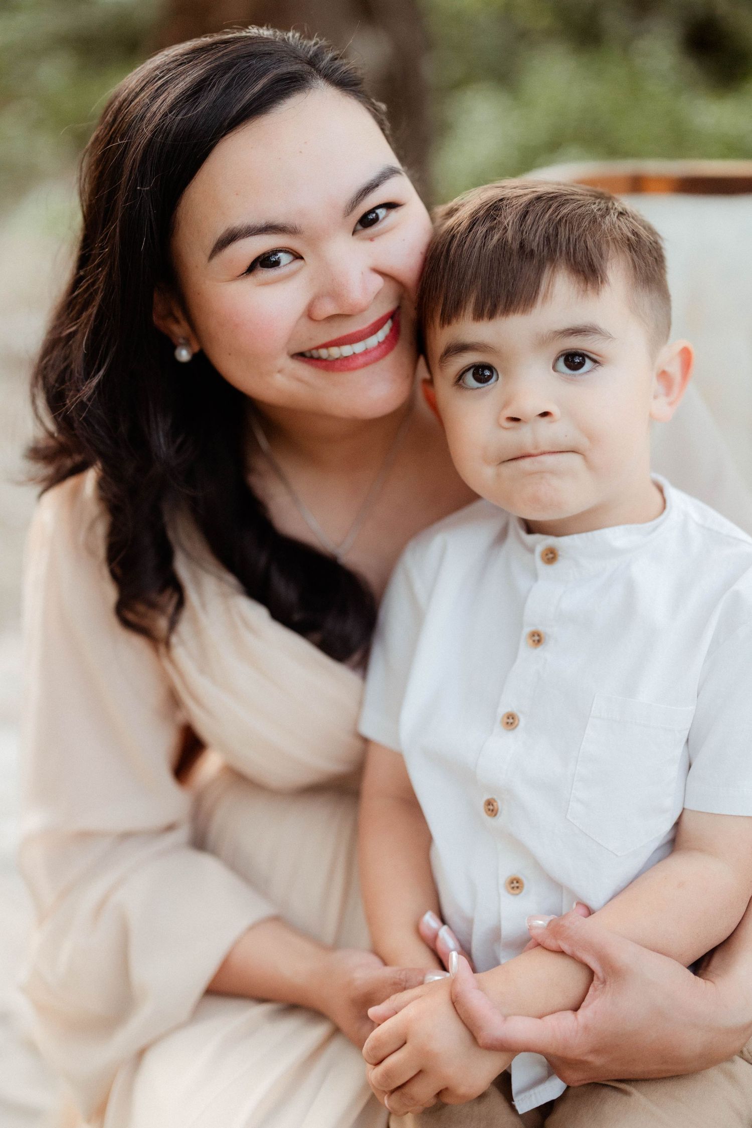 A smiling mother with dark hair hugs her young son in a white shirt outdoors.