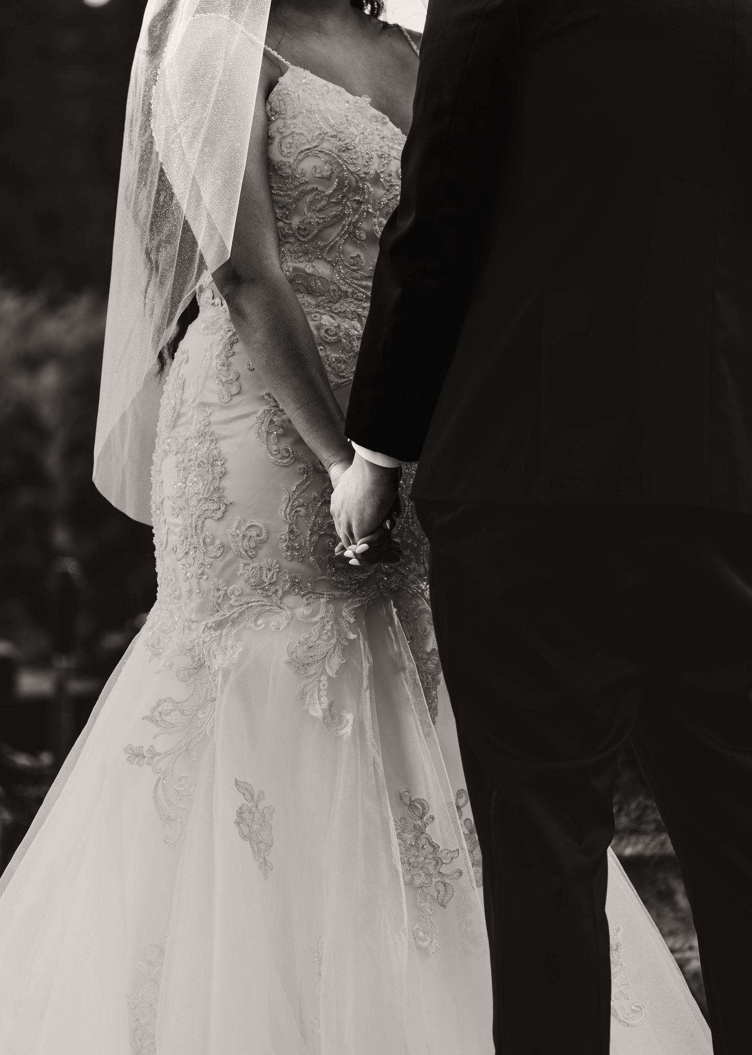 Black and white close-up of a bride in a lace mermaid gown holding hands with her groom.