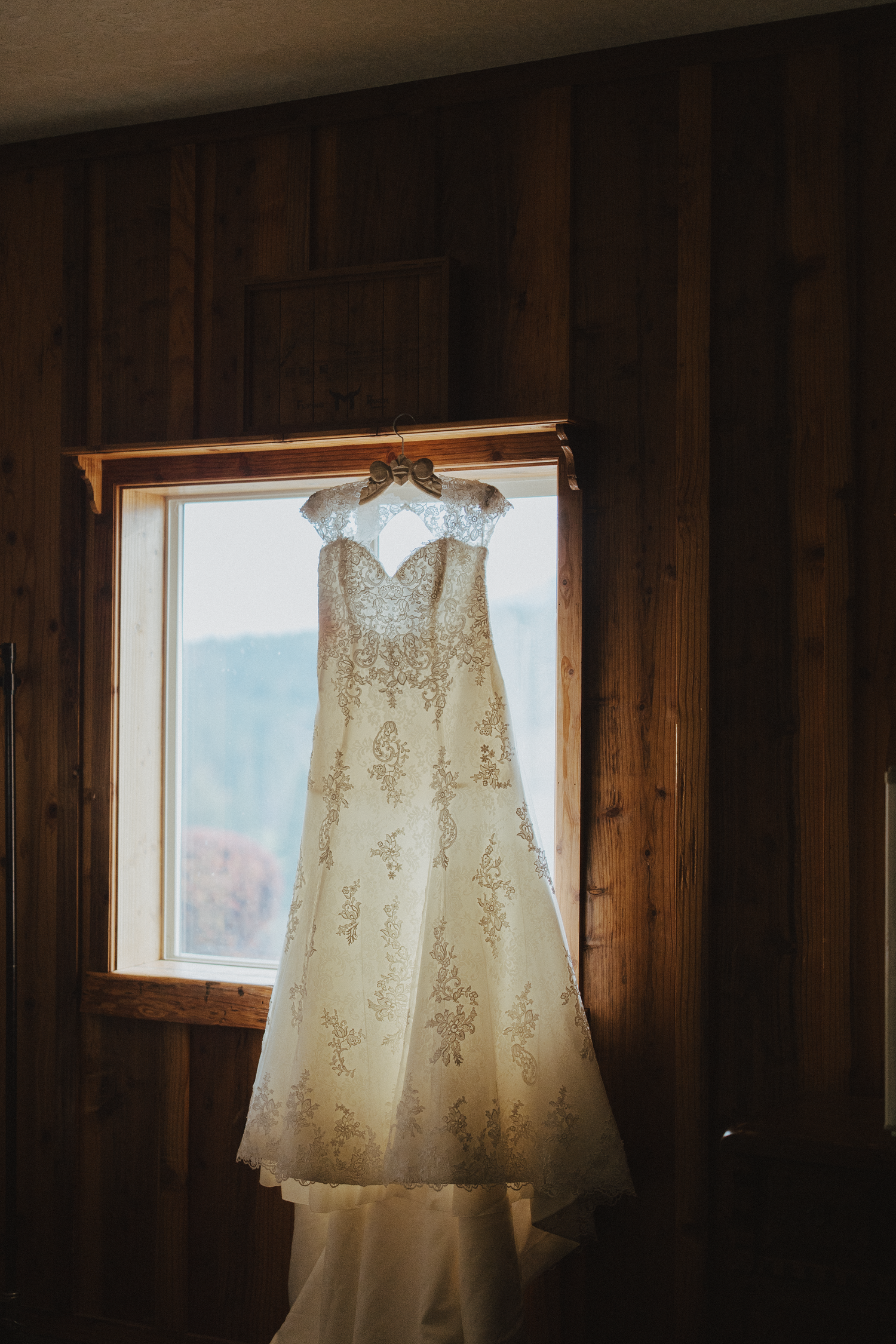 Elegant lace wedding dress hanging on hanger in front of rustic wooden window.