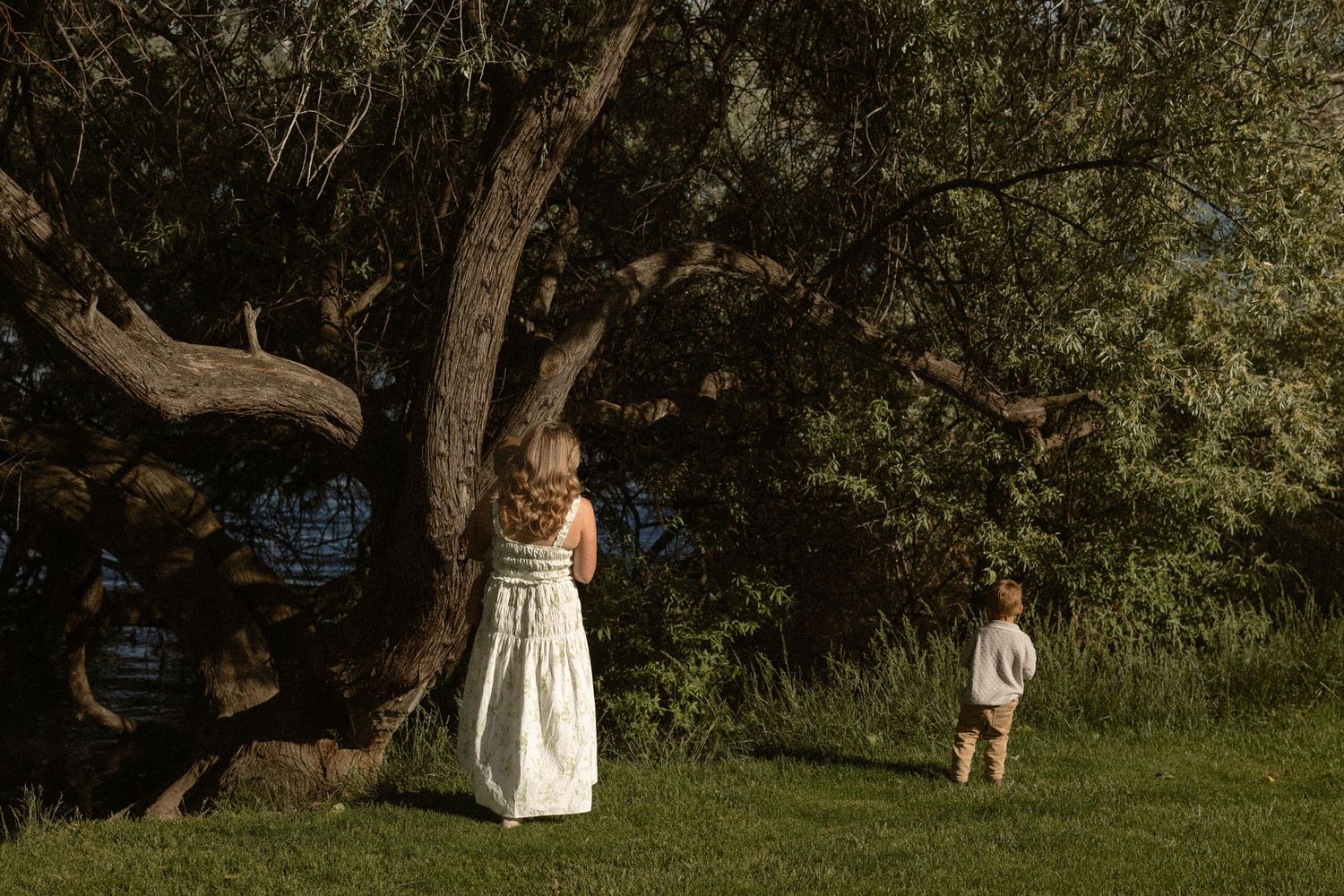 Mother in white dress and young child stand near large tree in lush green park.