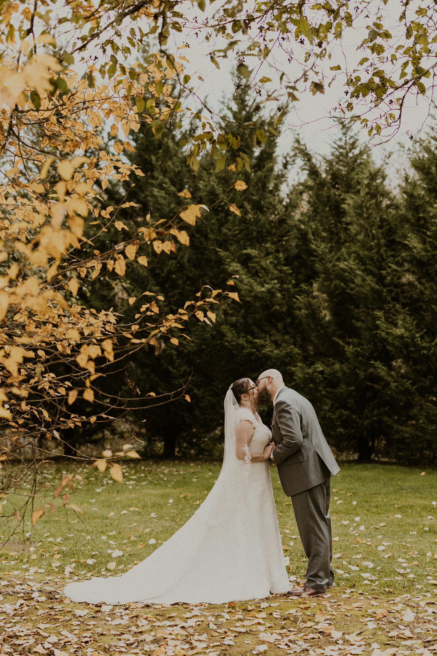 Bride and groom kissing under golden autumn leaves in a lush green park.