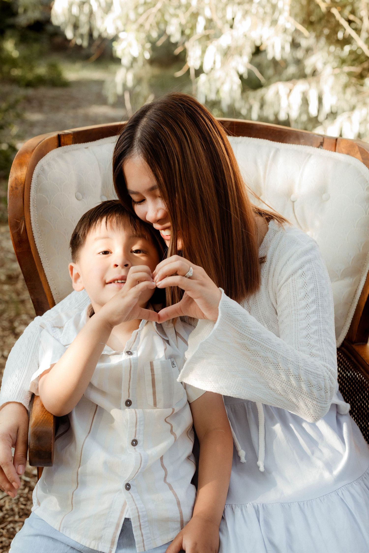 A smiling mother and her young son make a heart shape with their hands while sitting together in a white chair outdoors.