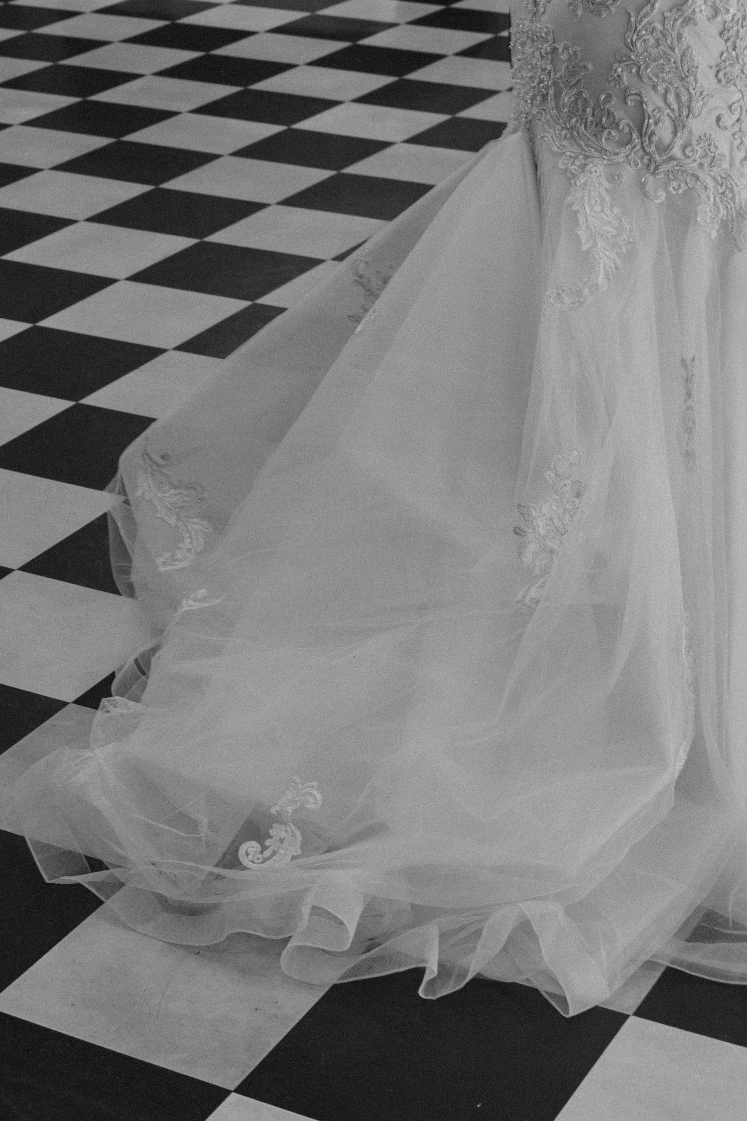 Close-up black and white photo of a bride's lace wedding gown train on a checkered tile floor.