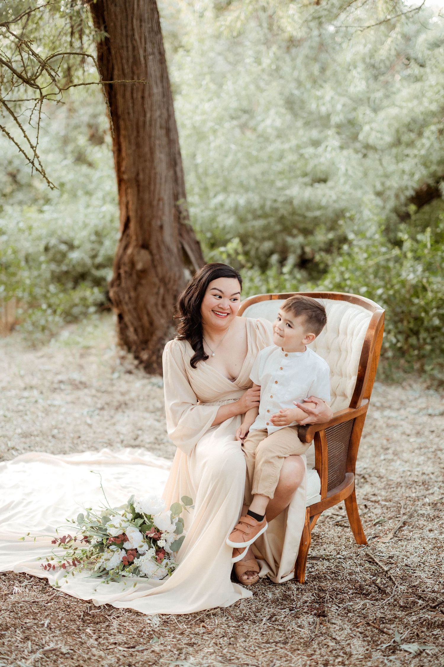 A woman in a beige dress sits with her young son on a wooden chair in a forest setting.