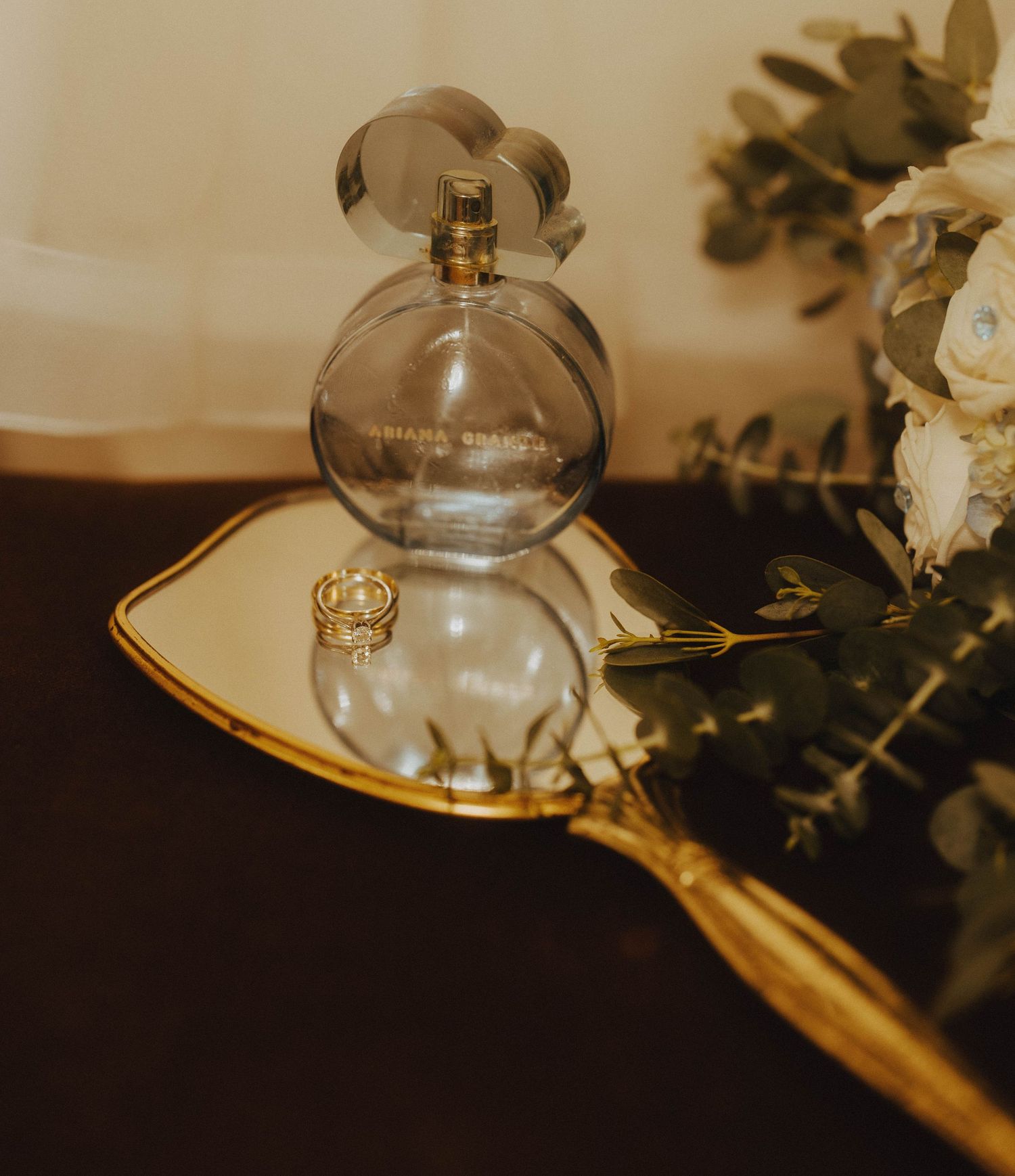 Elegant wedding flat lay featuring a Marc Jacobs perfume bottle, gold ring, and white flowers on a mirror tray.