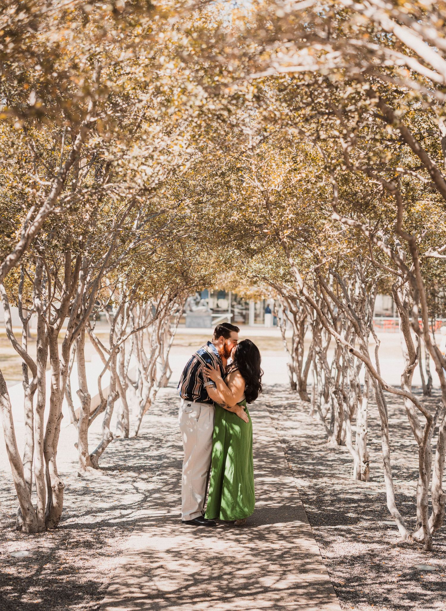 Couple sharing a romantic kiss under a canopy of trees in a sunlit park, woman wearing a green dress.