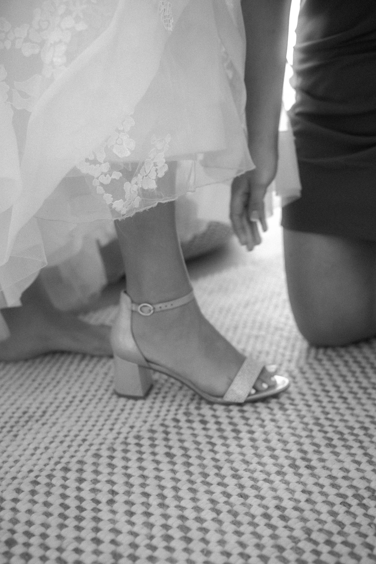 Black and white close-up of a bride having her strappy block-heel wedding shoe fastened by a bridesmaid.