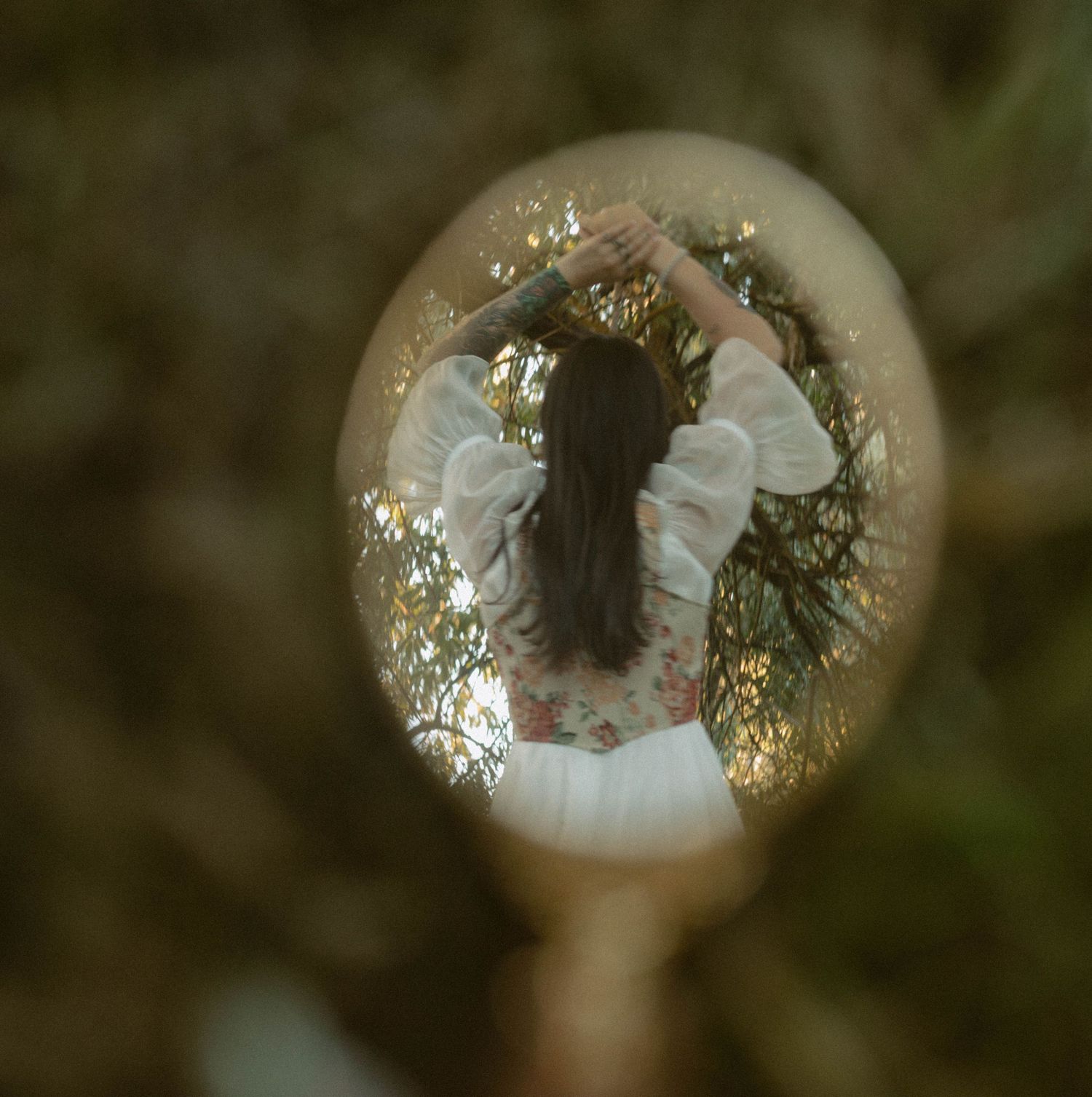 A girl in a white dress with floral details is reflected in a round mirror surrounded by lush green trees.