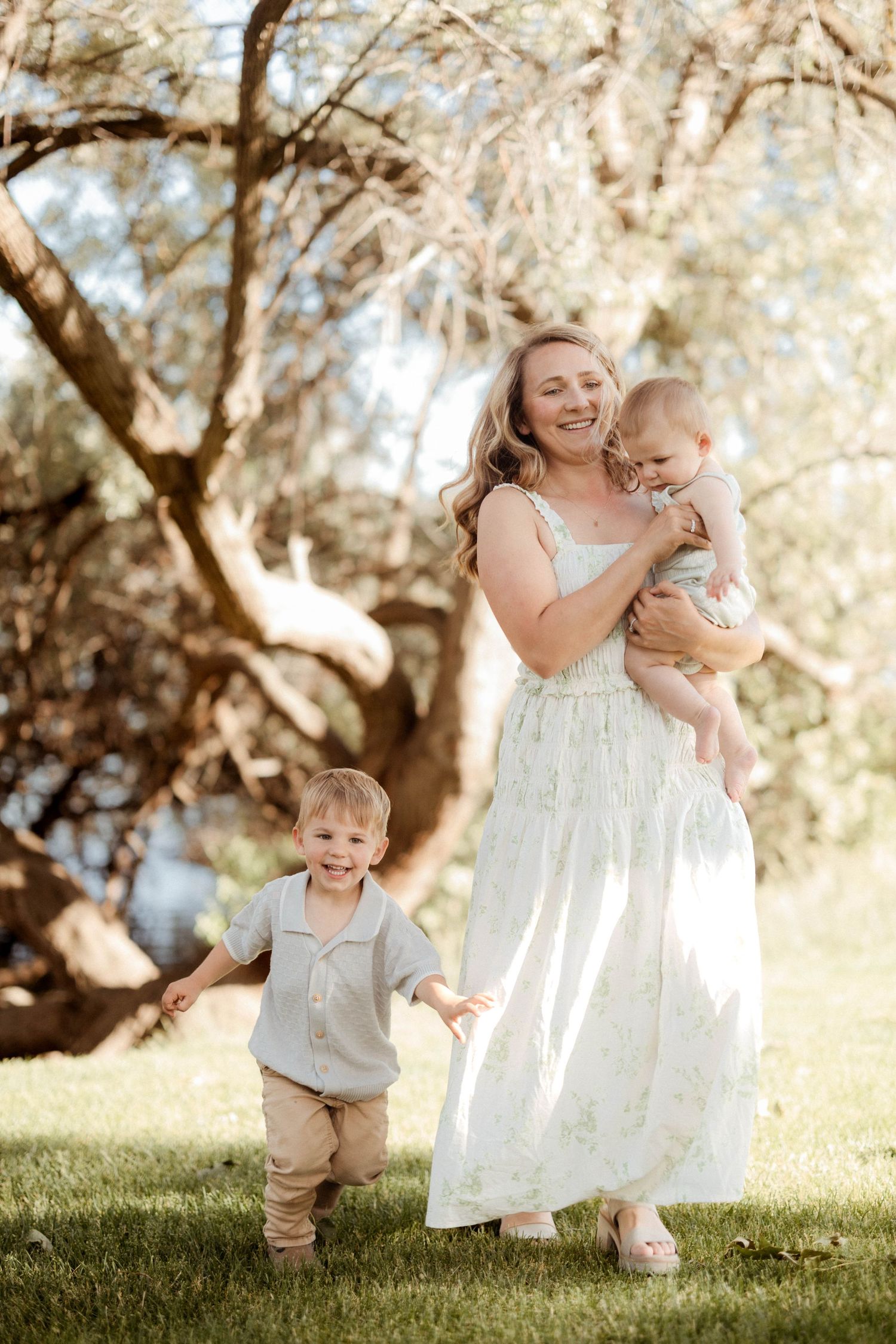 A smiling mother in a floral dress holds her baby while her toddler son walks beside her in a sunny park.