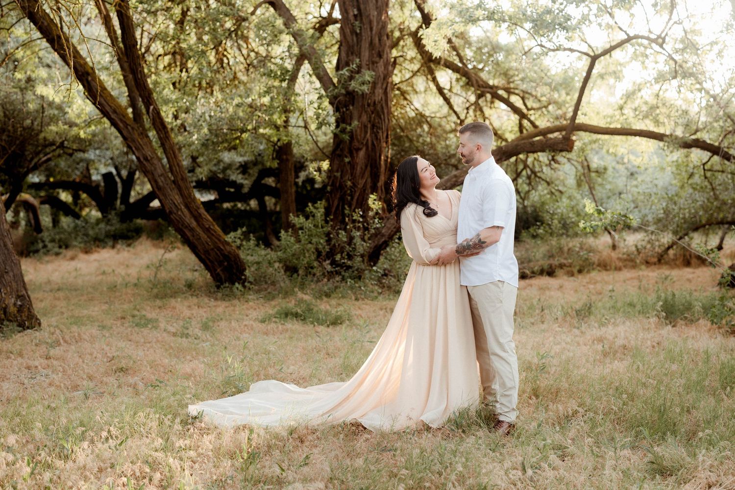 Couple sharing a tender moment in a sunlit meadow, woman wearing an elegant flowing champagne dress.
