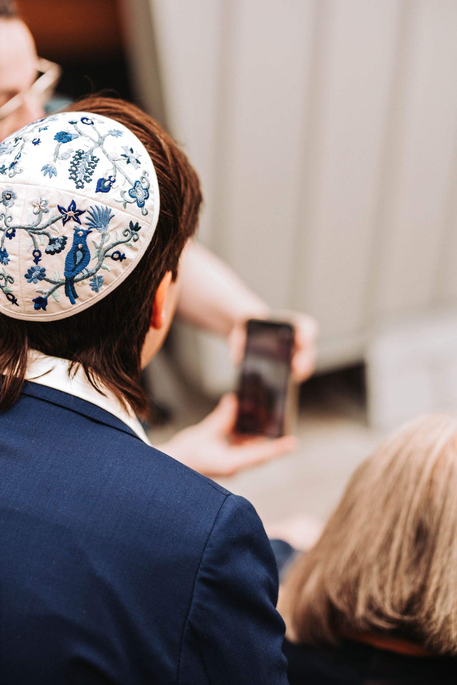 Man wearing an embroidered floral kippah at a Jewish wedding ceremony while someone holds a phone.