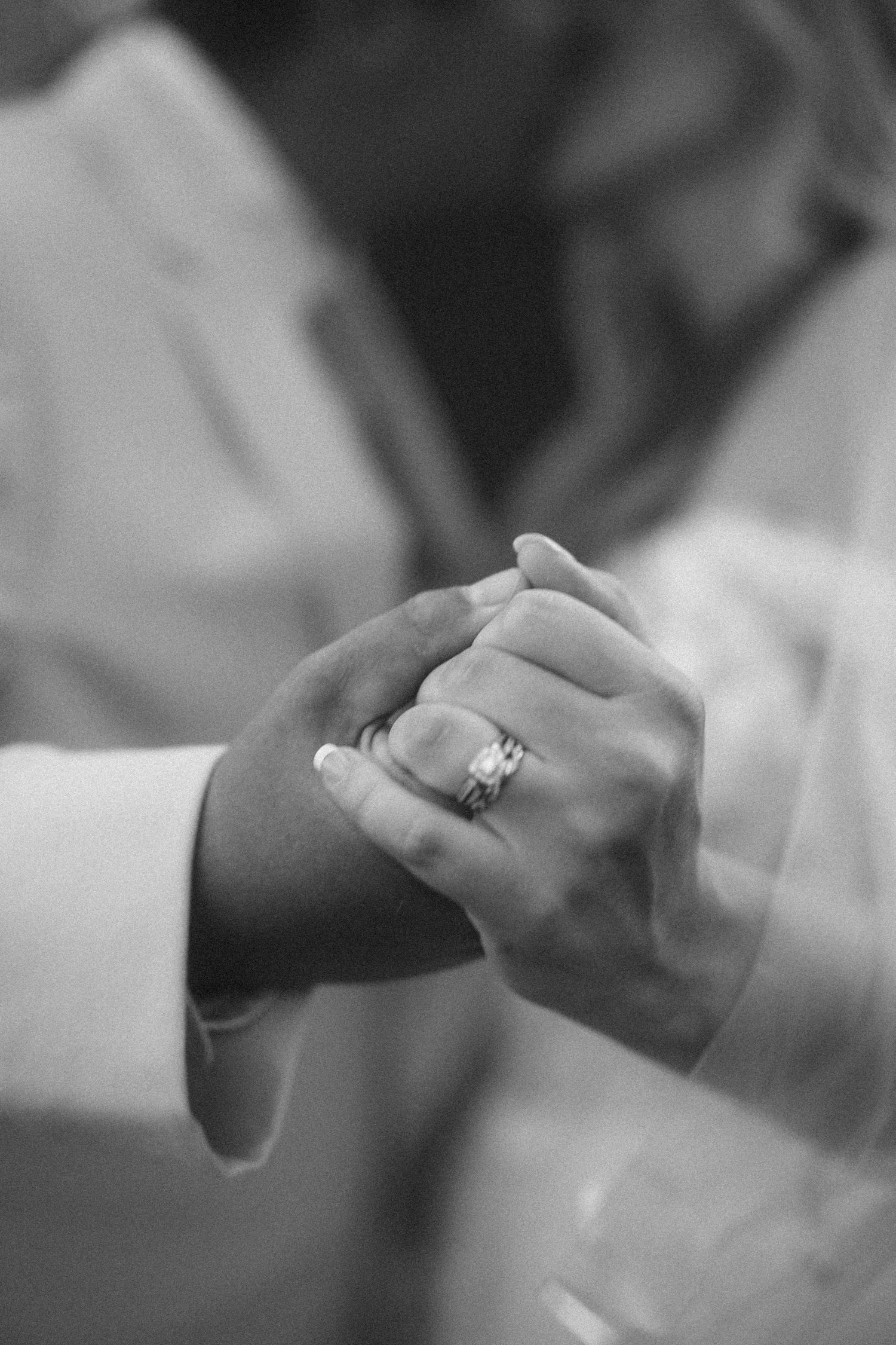 Black and white close-up of bride and groom holding hands, showcasing the bride's diamond wedding ring.