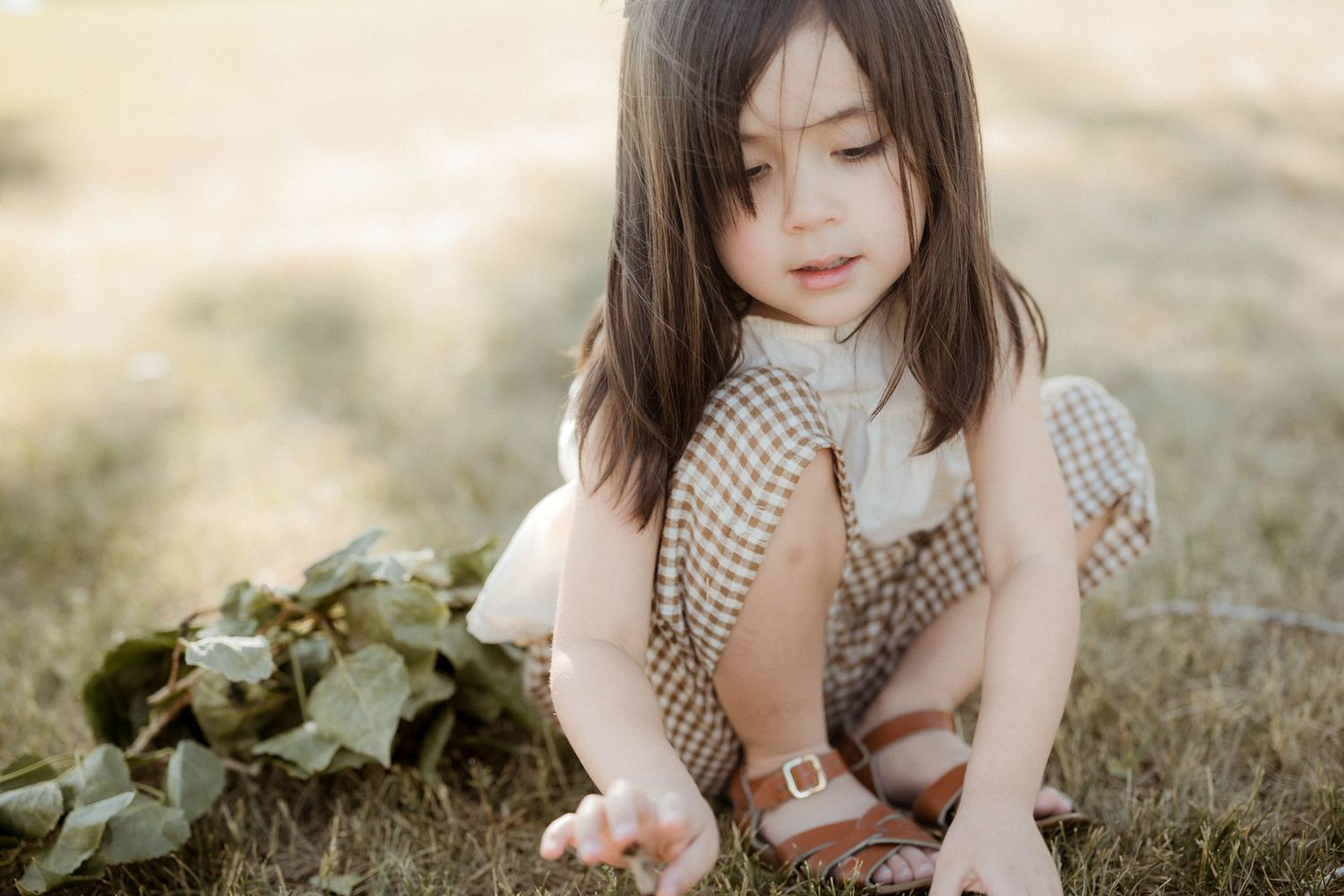 A young girl in a plaid romper sits on grass, looking down at her hand, with green leaves beside her.