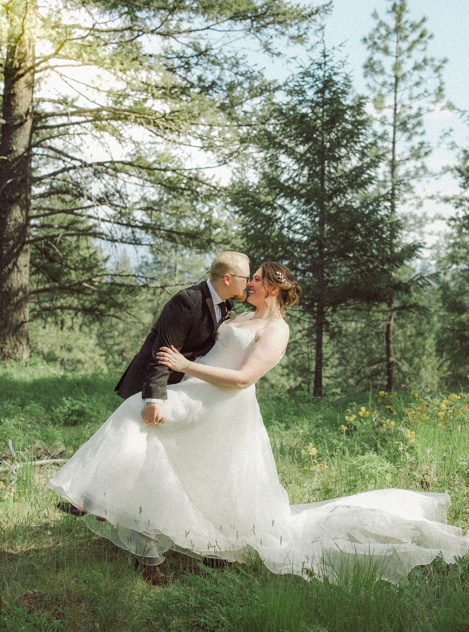 Groom dips bride for a romantic kiss in a sunny woodland clearing, her white gown trailing on the grass.
