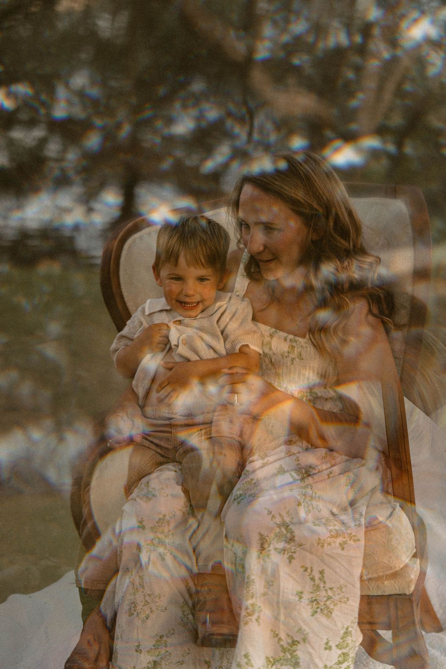 Mother and smiling toddler sit together in vintage chair in soft double-exposure outdoor photo.