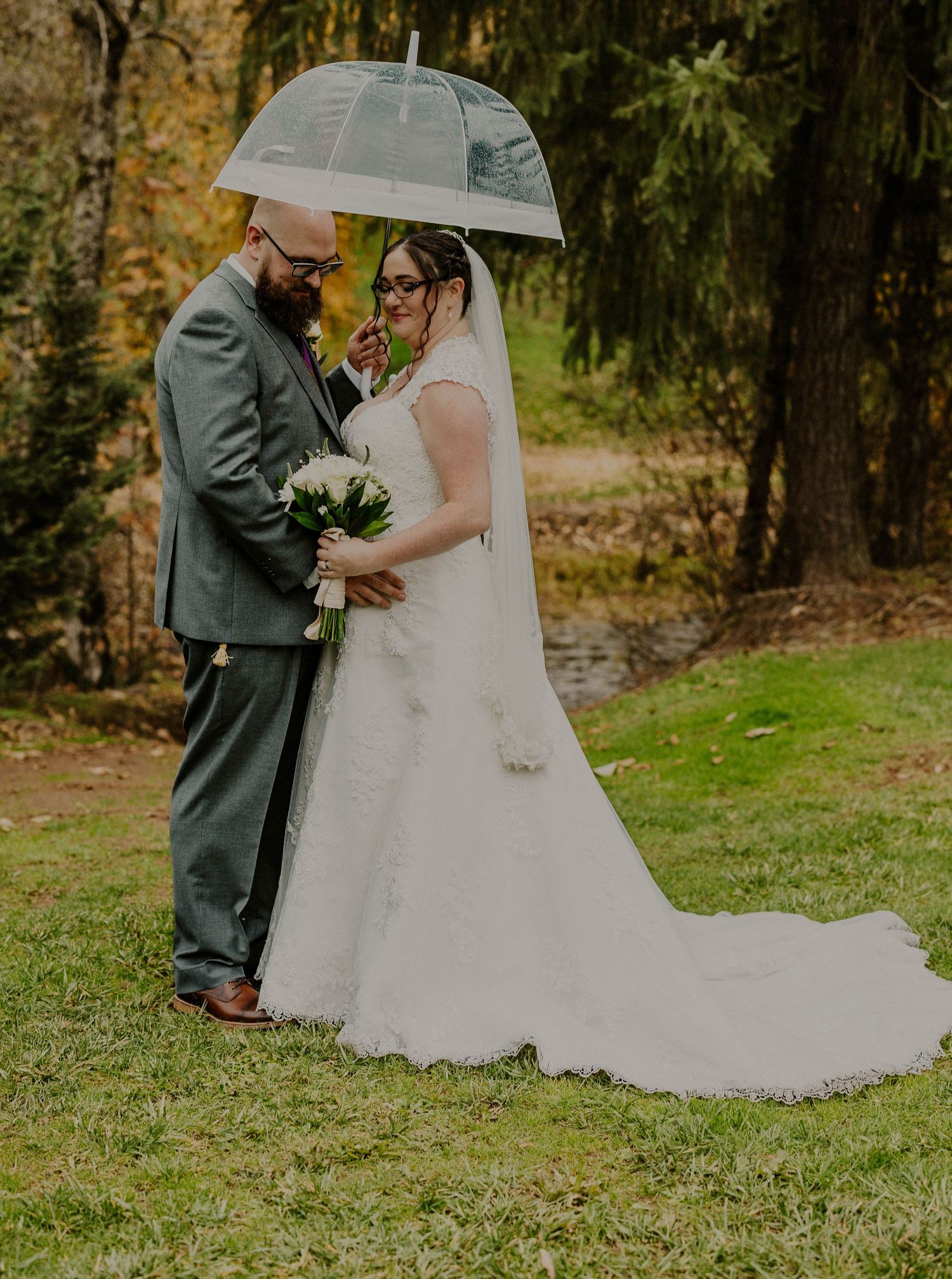 Bride and groom sharing intimate moment under clear umbrella on rainy wedding day.
