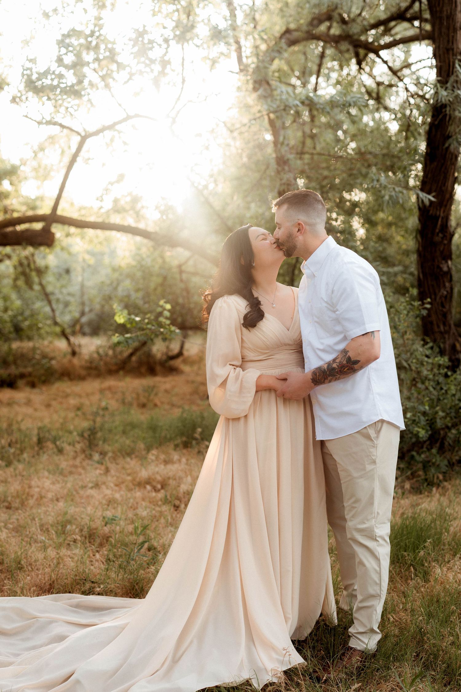Couple sharing a tender kiss in sun-flared woodland setting, woman in flowing champagne maternity gown.