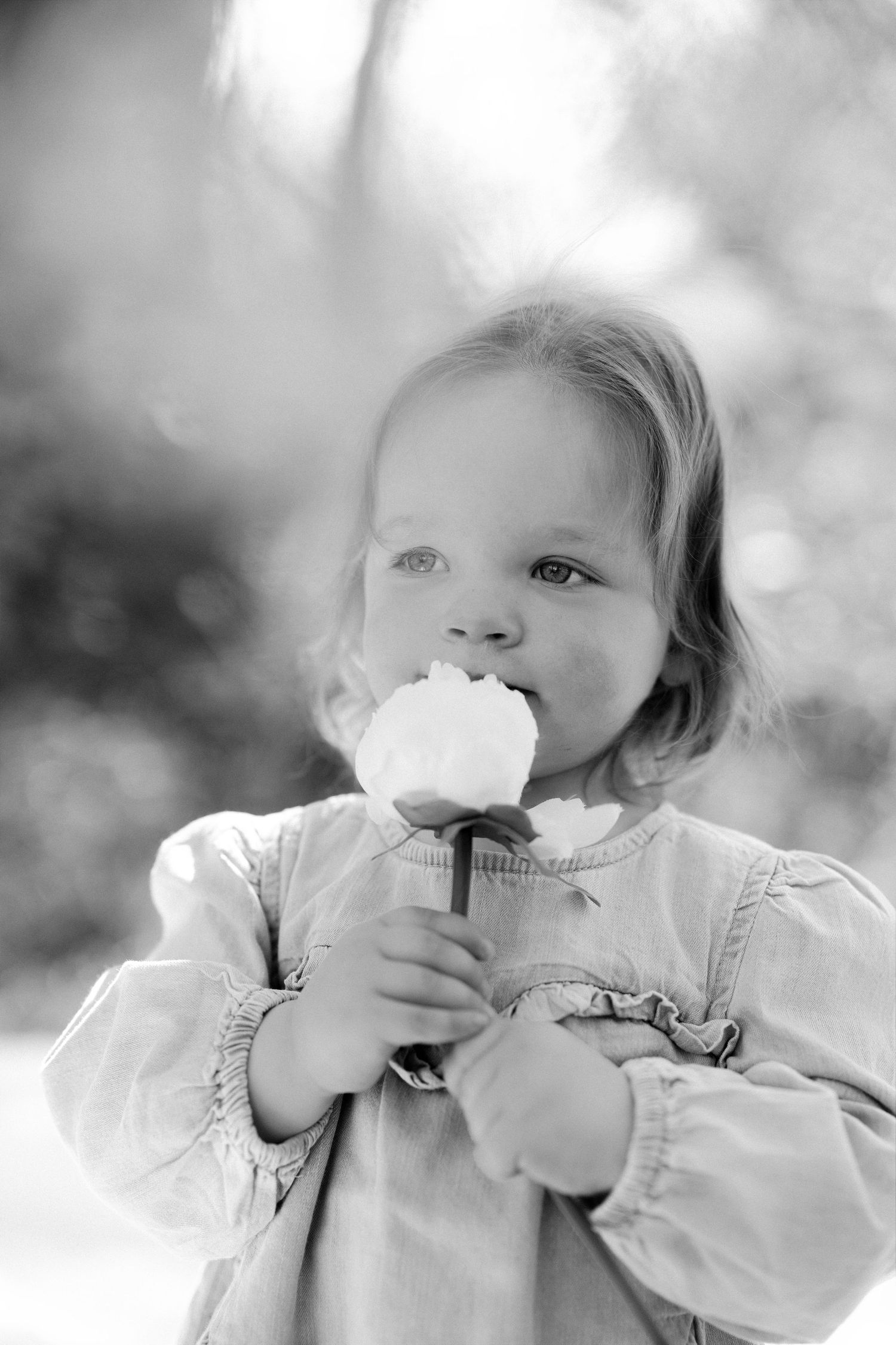 A toddler girl in a ruffled dress holds and smells a white flower outdoors in black and white.