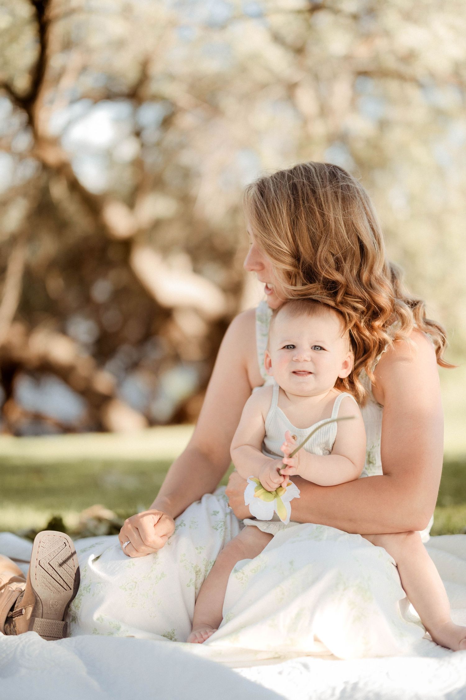A blonde mother in a floral dress sits outdoors on a blanket holding her smiling baby in soft natural light.
