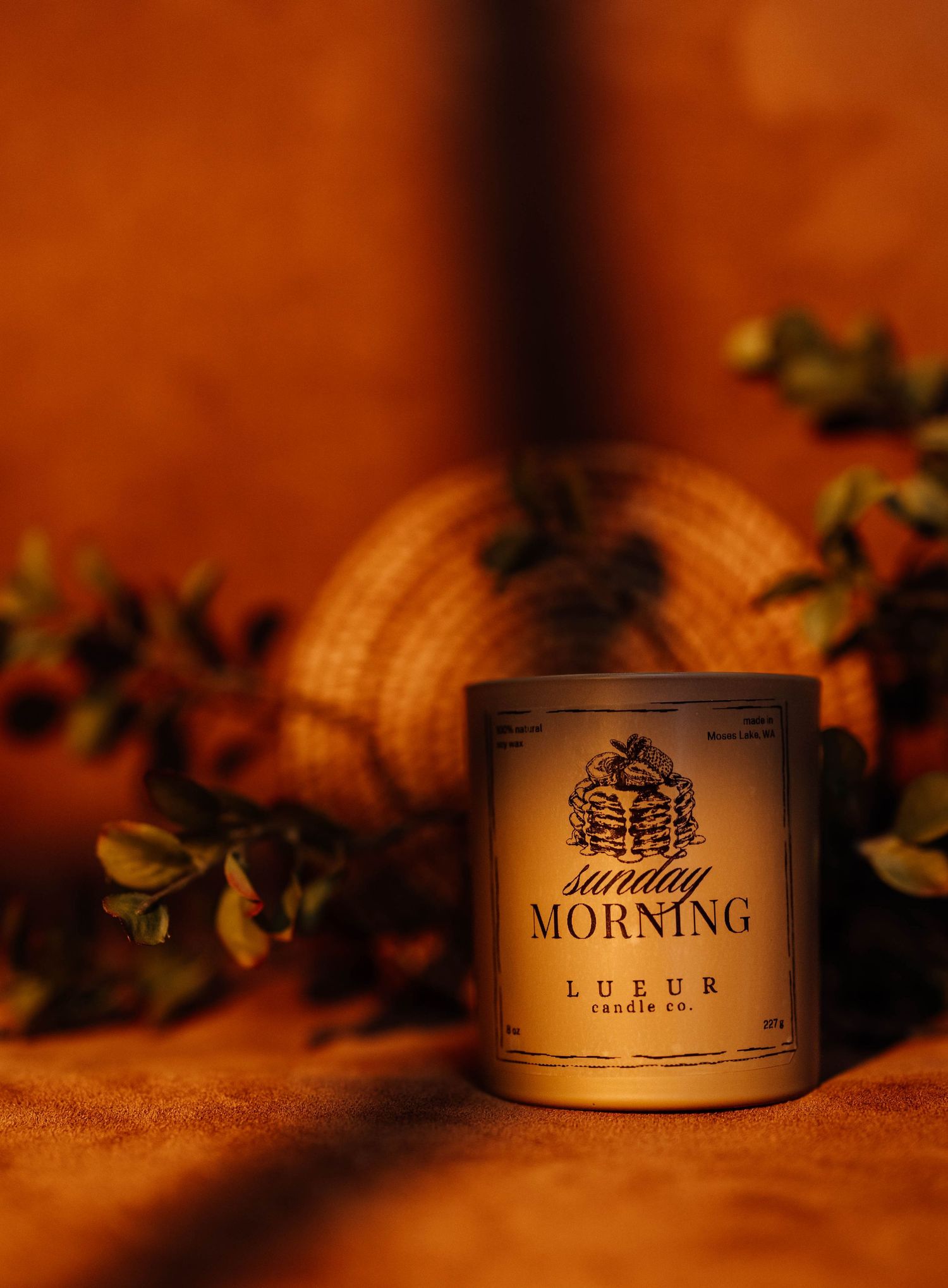 A Sunday Morning scented candle by Lueur Candle Co. displayed with autumn foliage on a warm amber background.