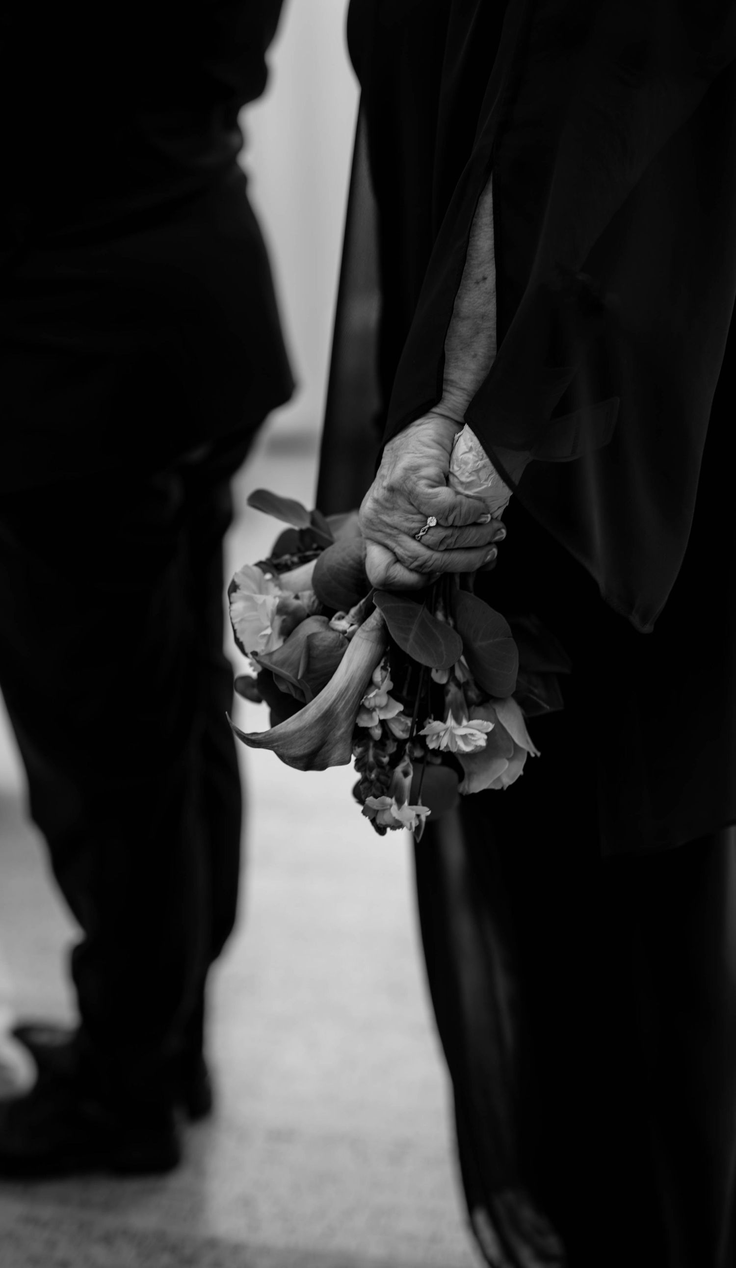 Black and white close-up of an elderly couple holding hands with a small floral bouquet.