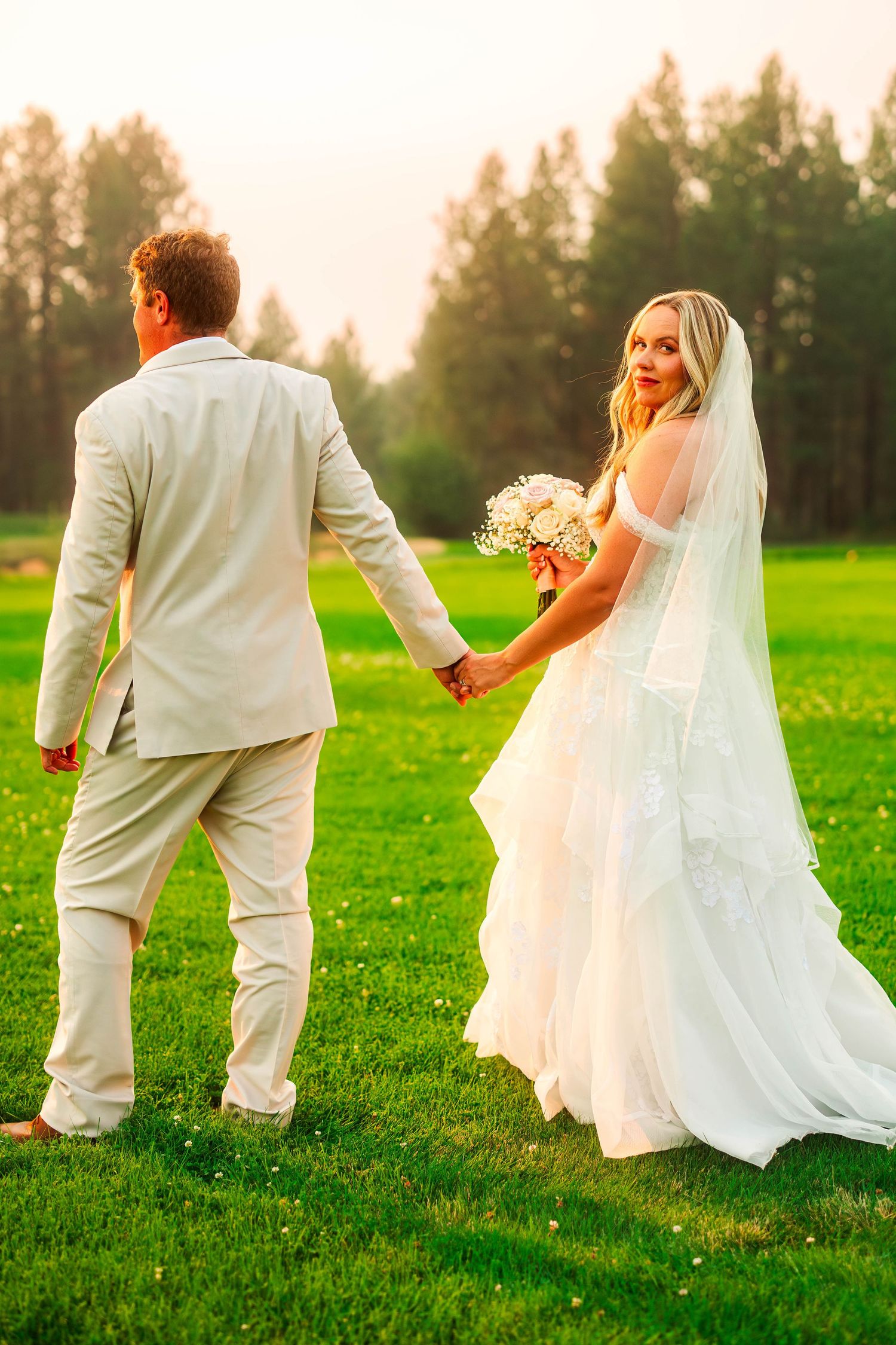 Bride in a white gown holding a bouquet looks back while holding hands with groom at golden hour.