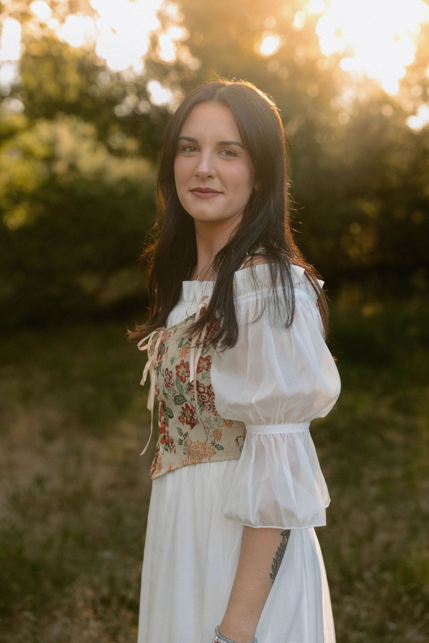 Woman in white embroidered dress standing in golden sunlight outdoors.