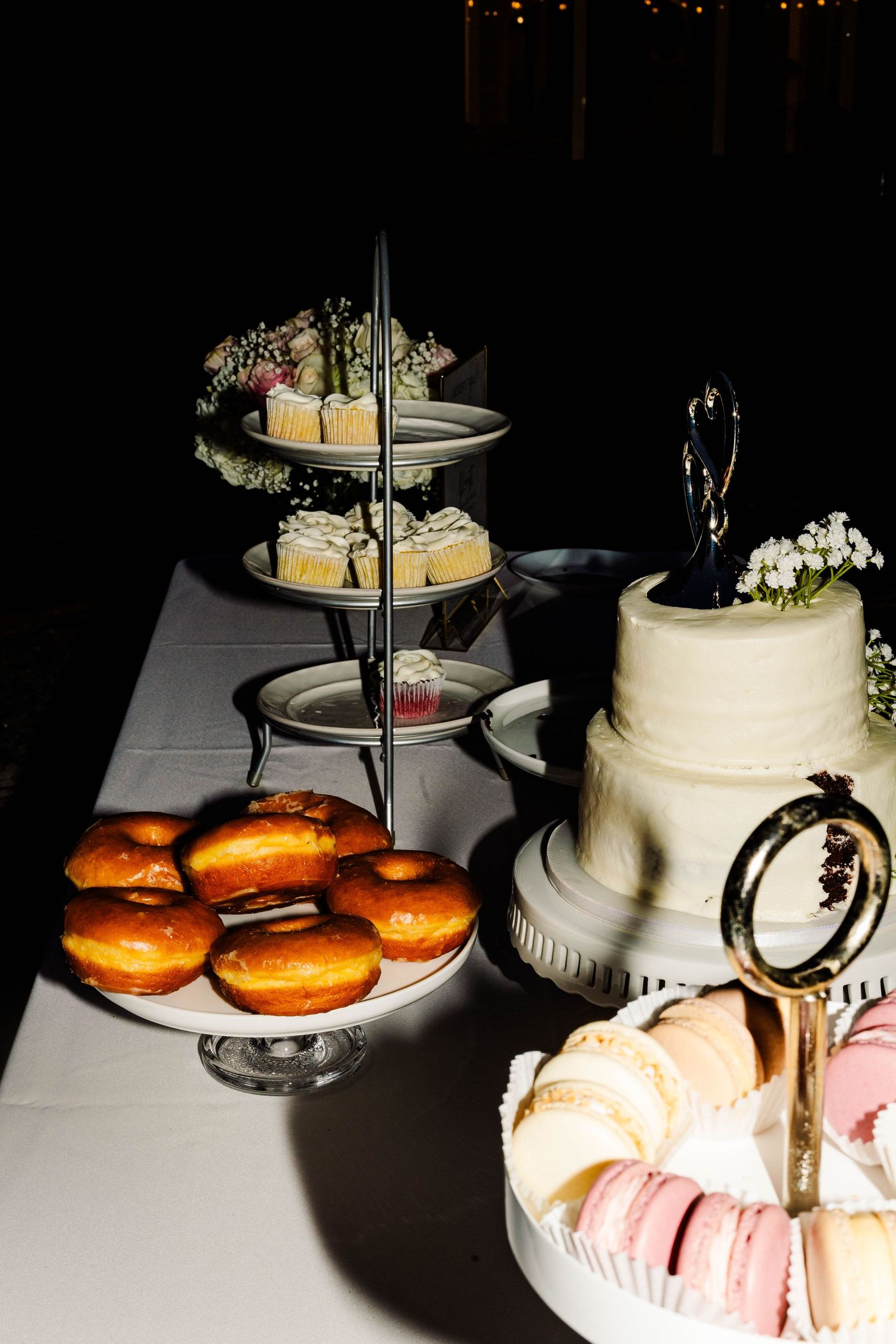 Wedding dessert table featuring white tiered cake, glazed donuts, cupcakes, and colorful macarons at night.