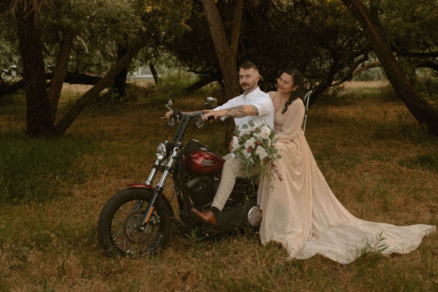 Bride and groom laughing together on a vintage red motorcycle surrounded by autumn trees.