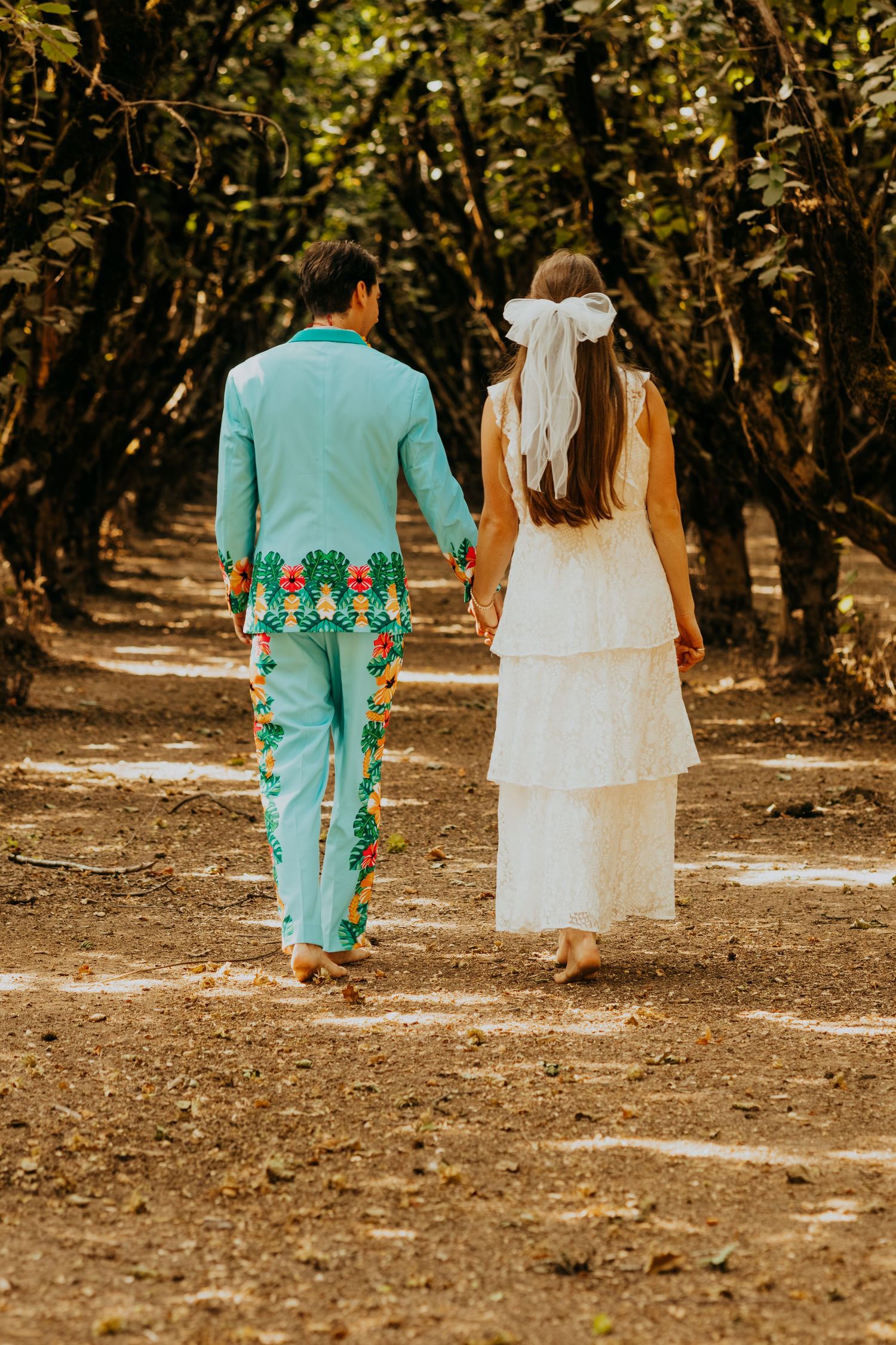 Bride and groom walk hand in hand down a tree-lined path, groom wearing a vibrant tropical patterned suit.