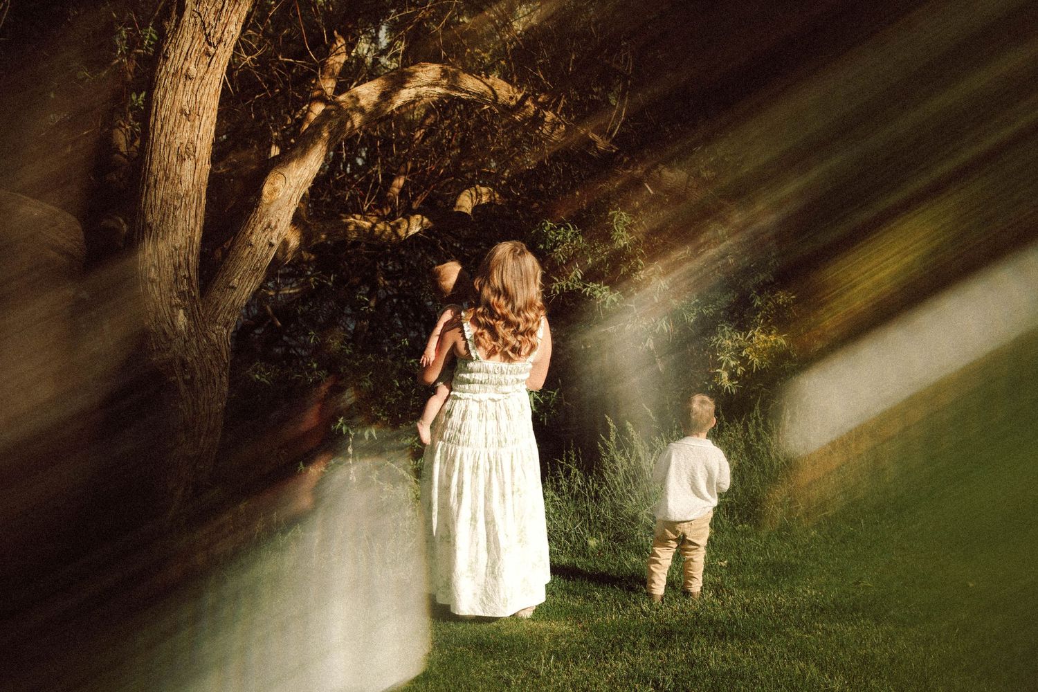 A mother in a white dress holds her child near a tree while a young boy stands on green grass in warm evening light.