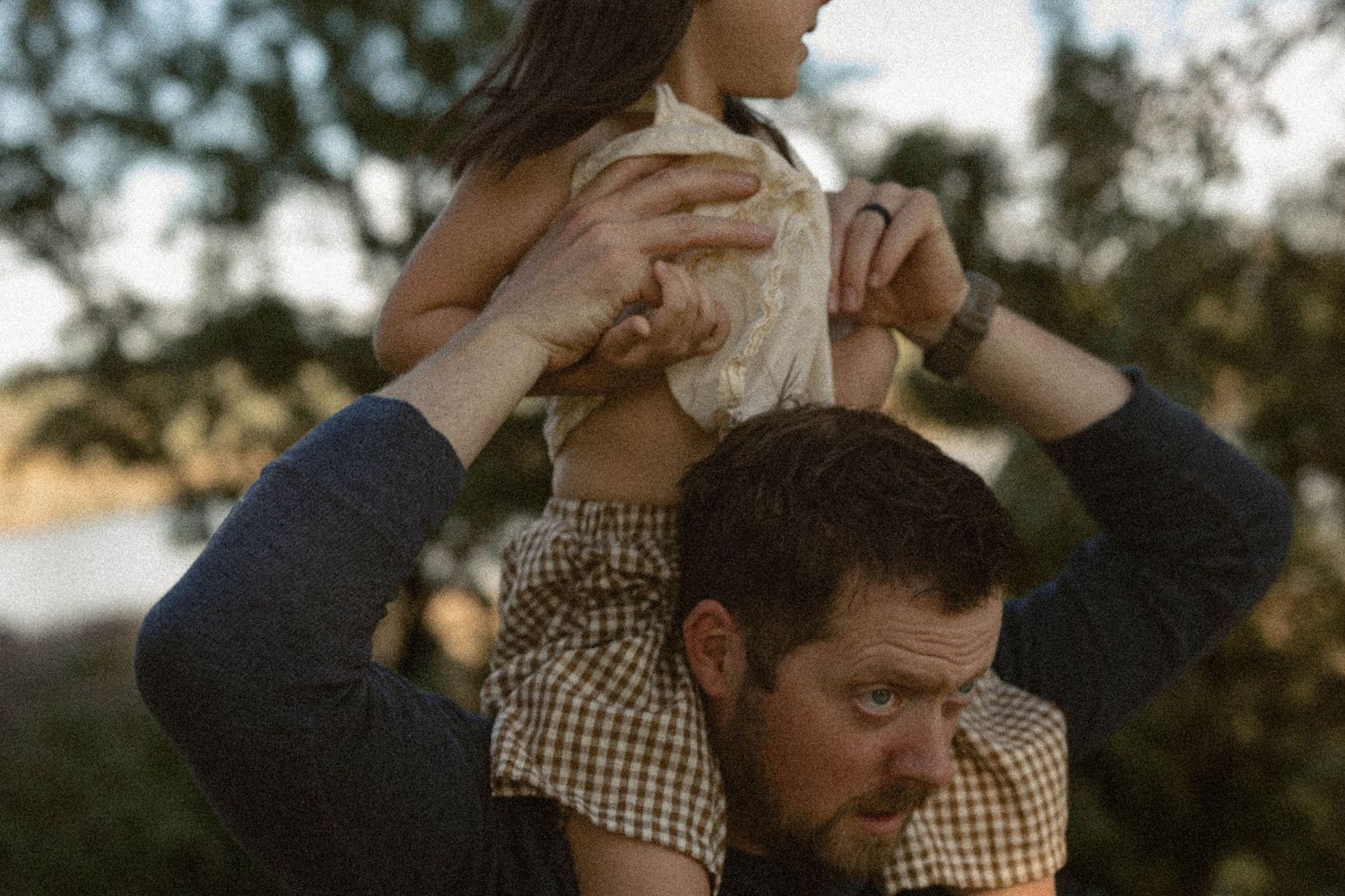 A father lifts his young daughter onto his shoulders outdoors with trees and sky in the background.