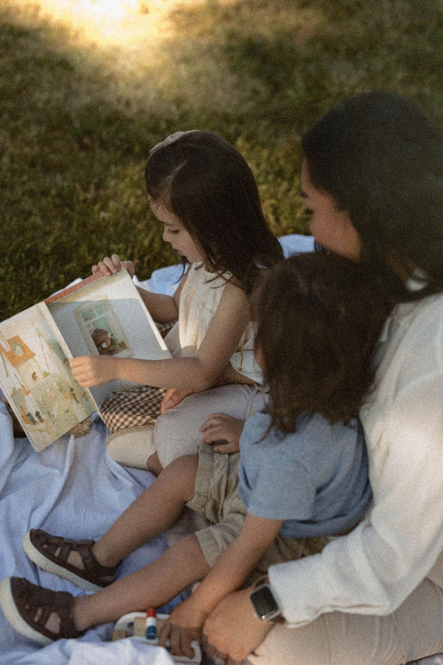 Mother reads a picture book with two young children on a blanket in a sunlit park.
