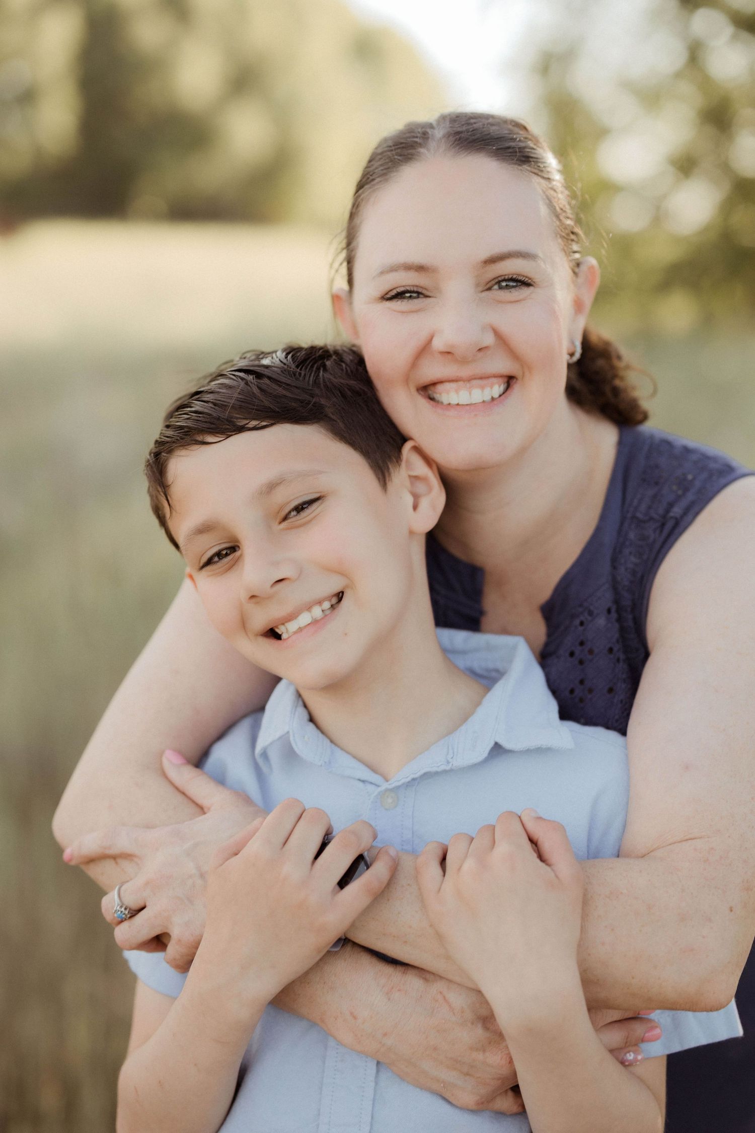 A smiling mother hugs her young son from behind in a sunny outdoor field setting.