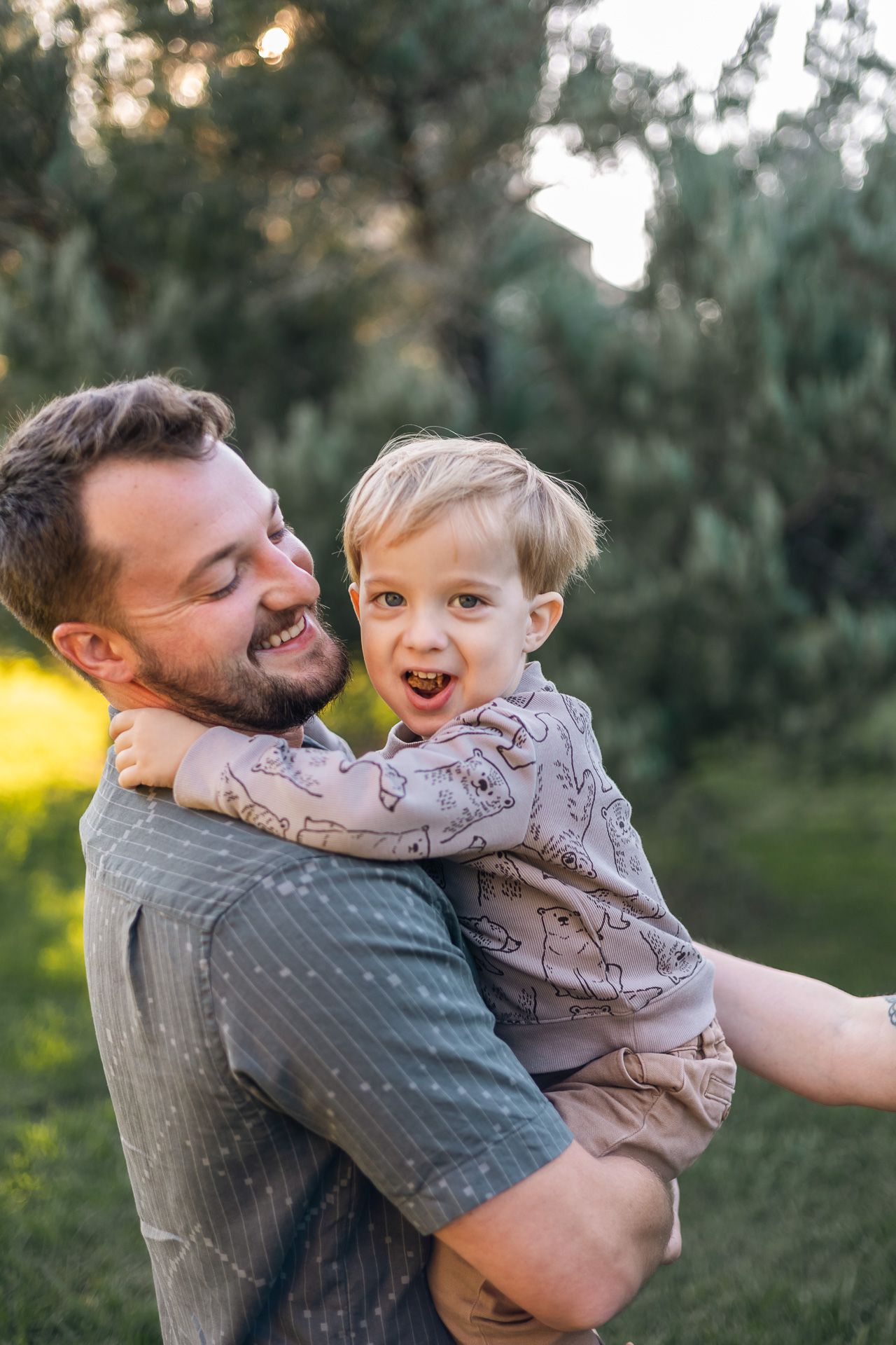 Smiling bearded father holds laughing toddler son outdoors in lush green garden at sunset.