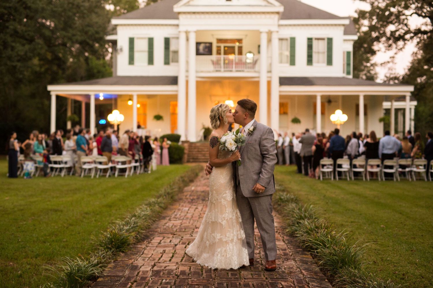 Bride and groom kissing on a brick path in front of a white plantation-style mansion at dusk.