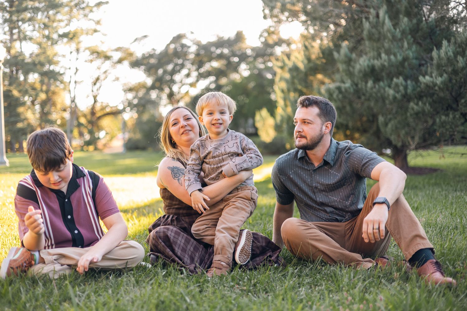 Family of four relaxes together on green grass in sunny park during warm golden afternoon light.