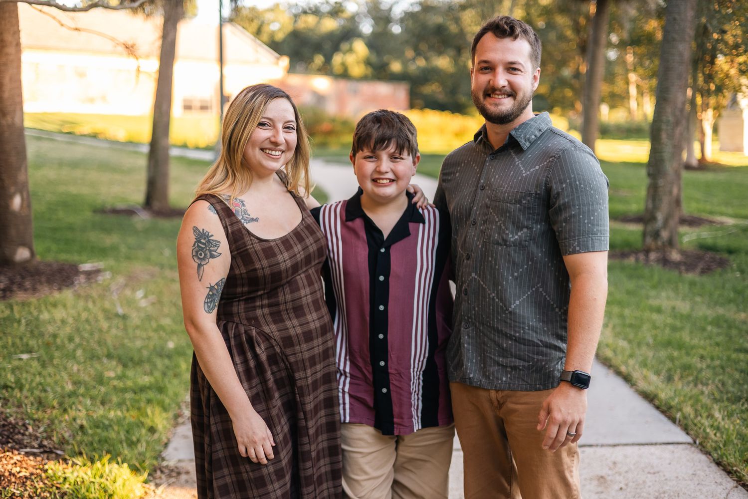 Smiling family of three poses together on sunny sidewalk path surrounded by green trees and lawn.