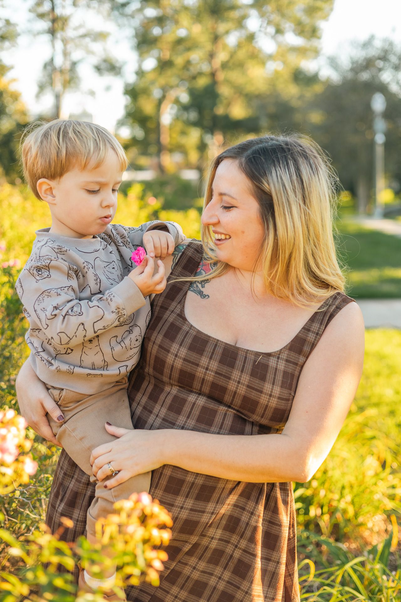 Smiling mother holds curious toddler boy who examines pink flower in sunlit garden setting.